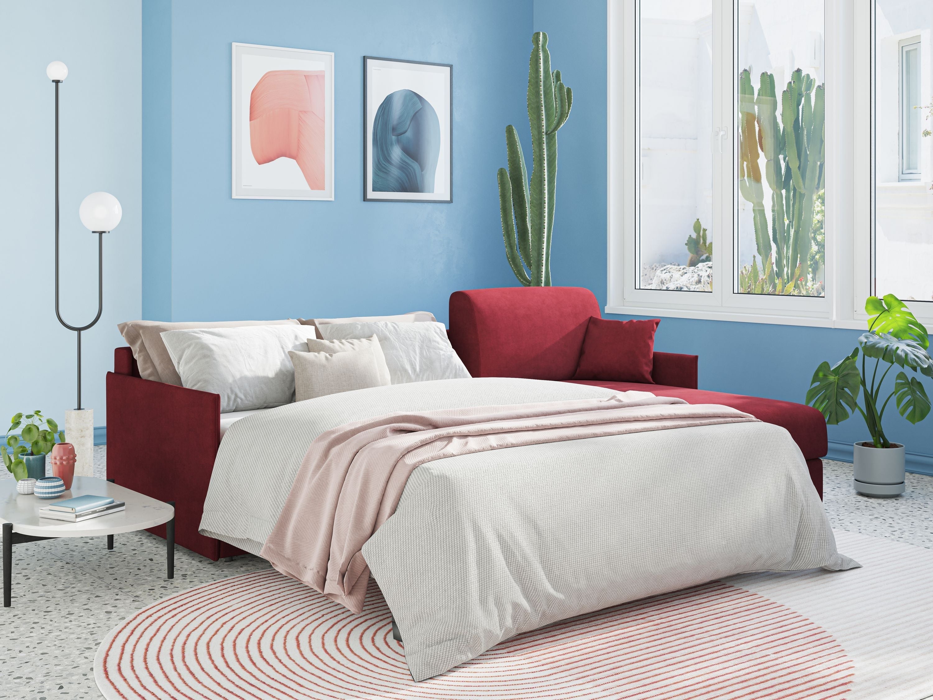 Una camera da letto moderna con un letto rosso, lenzuola bianche e rosa, pareti blu, arte astratta, grandi finestre, cactus alti, piante verdi, un tappeto rotondo a strisce rosse e un piccolo tavolino con una lampada e dei libri.