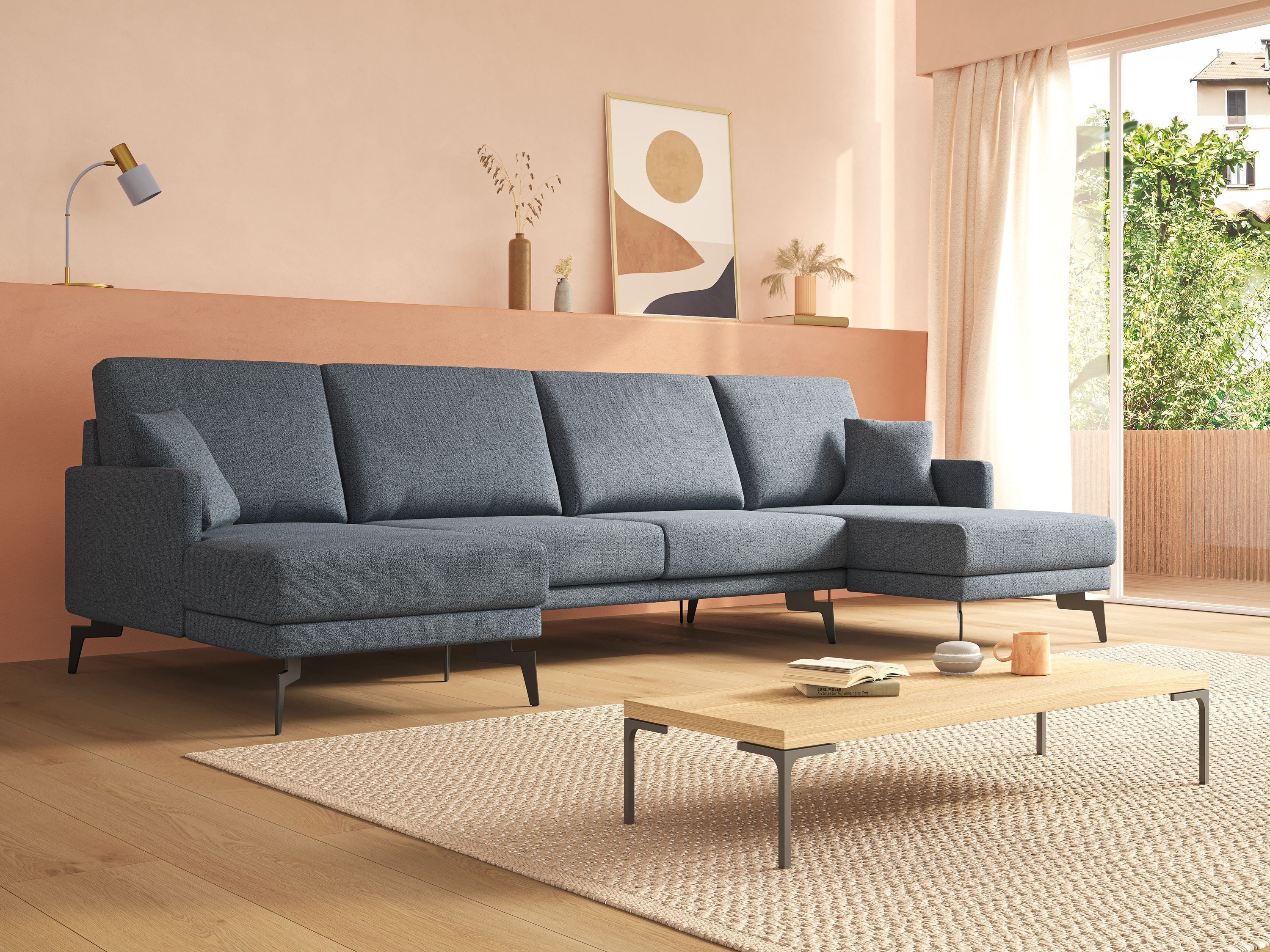 Un soggiorno moderno con un grande divano componibile blu, un tavolino da caffè in legno chiaro, un tappeto beige, quadri astratti alle pareti e una grande finestra che lascia entrare la luce naturale, con vegetazione visibile all'esterno.