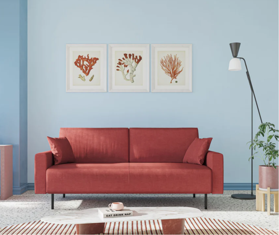 Un soggiorno moderno con un divano rosso, due cuscini rossi, un tavolino da caffè bianco, una lampada da terra a treppiede, una pianta in vaso e tre stampe botaniche incorniciate su una parete azzurra.