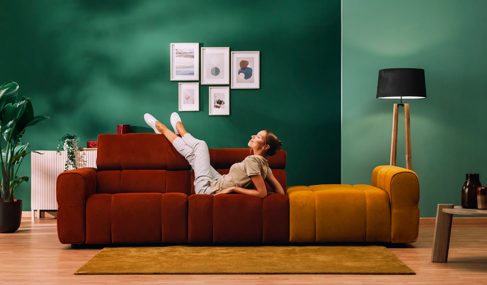 Una donna è sdraiata con le gambe sollevate su un divano color ruggine e senape in un soggiorno moderno con pareti verdi, quadri incorniciati, una lampada da terra, una pianta e un tappeto marrone chiaro su un pavimento in legno.