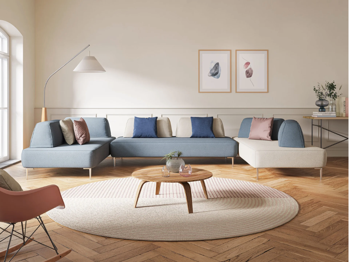 Soggiorno moderno con divano componibile azzurro e beige, cuscini assortiti, tavolino rotondo in legno su un tappeto circolare, quadri alla parete, lampada da terra, tavolino laterale con vaso e grande finestra con luce naturale.