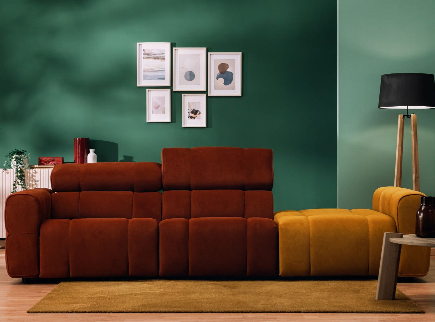 Un soggiorno moderno con un divano componibile color ruggine e senape, una parete decorativa verde, un quadro con cornice, una lampada da terra nera, un tavolo in legno e un tappeto color senape su un pavimento in legno chiaro.