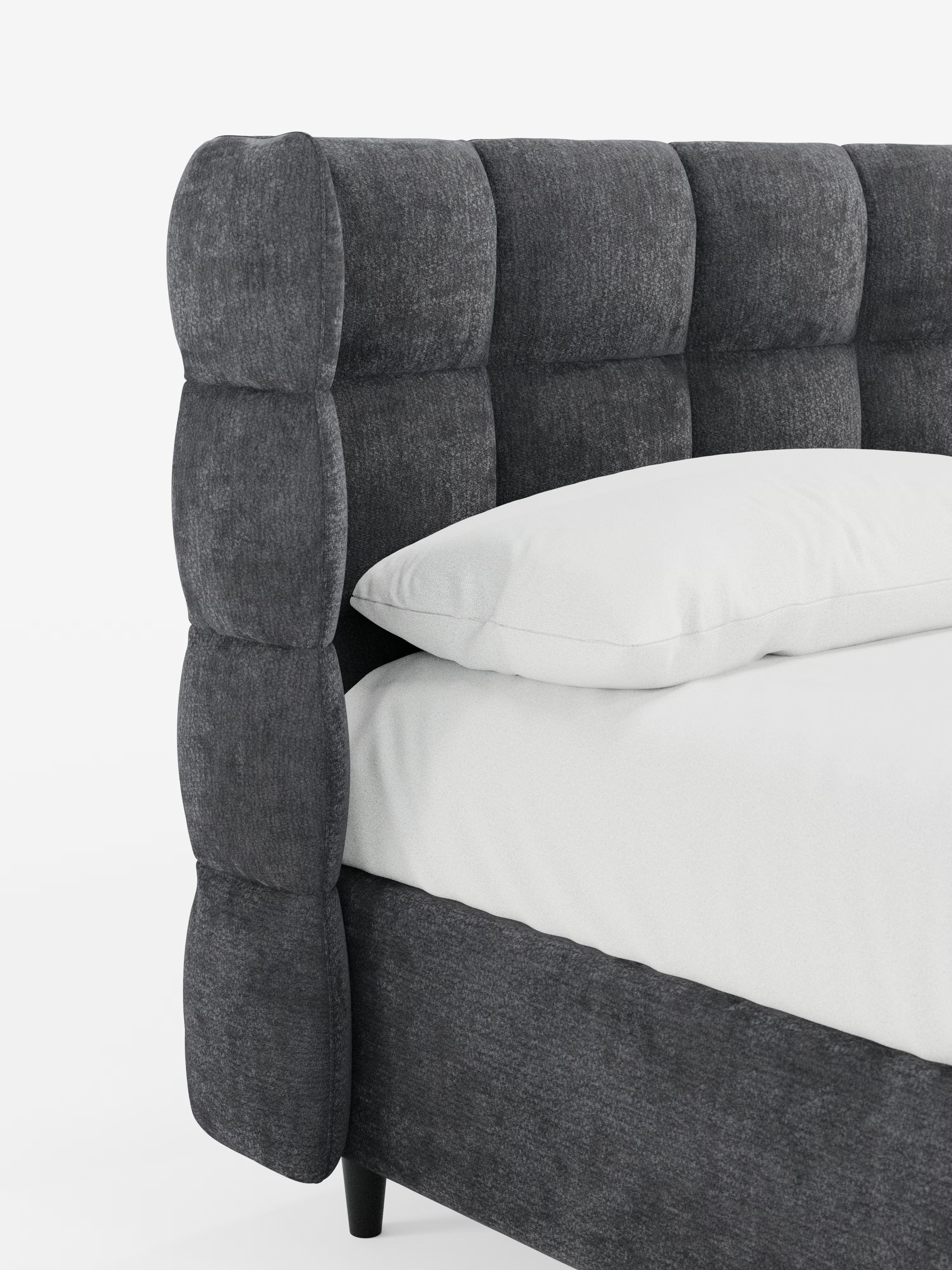 Primo piano del letto imbottito RAGNAR con contenitore e testiera capitonné avvolgente di Letti.store, caratterizzato da una testiera imbottita grigio scuro con spesse e morbide sezioni quadrate in classico stile capitonné. Un elegante tessuto certificato Oeko-Tex® riveste sia la struttura del letto che la testiera abbinate. Sul letto, su uno sfondo bianco a tinta unita, poggiano un cuscino bianco e un lenzuolo bianco.