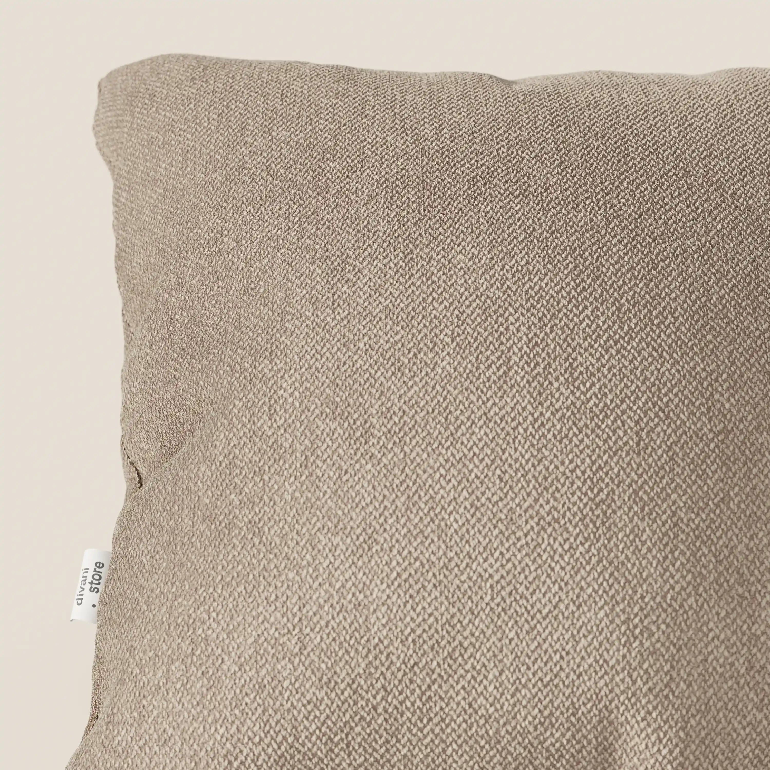 Primo piano dell'angolo superiore di un cuscino d'arredo beige Sogno di Divani.Store in morbido tessuto smacchiabile T05, che mostra la trama a spina di pesce e un'etichetta bianca cucita. Lo sfondo è semplice e di colore chiaro.