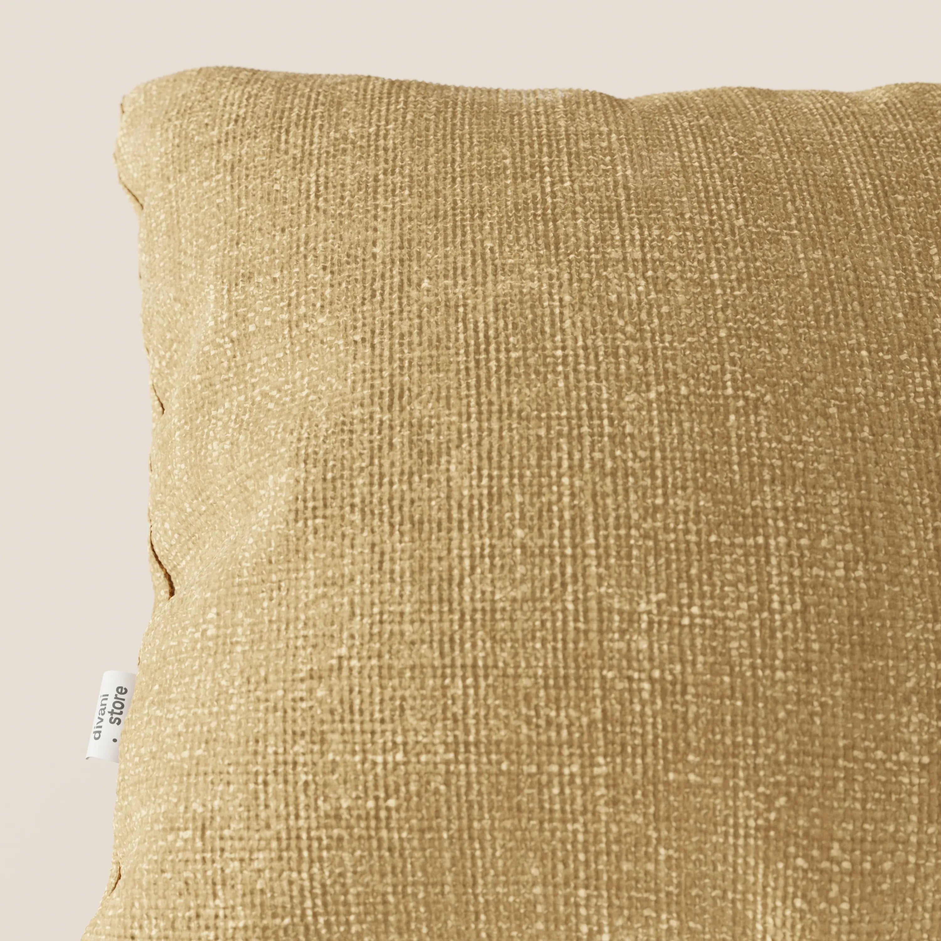 Primo piano del cuscino Lyon 45x45 cm di Divani.Store in T06 giallo, realizzato in morbido tessuto impermeabile. Su uno sfondo beige chiaro si vede solo una parte del cuscino giallo oro, con una piccola etichetta bianca sul bordo sinistro.