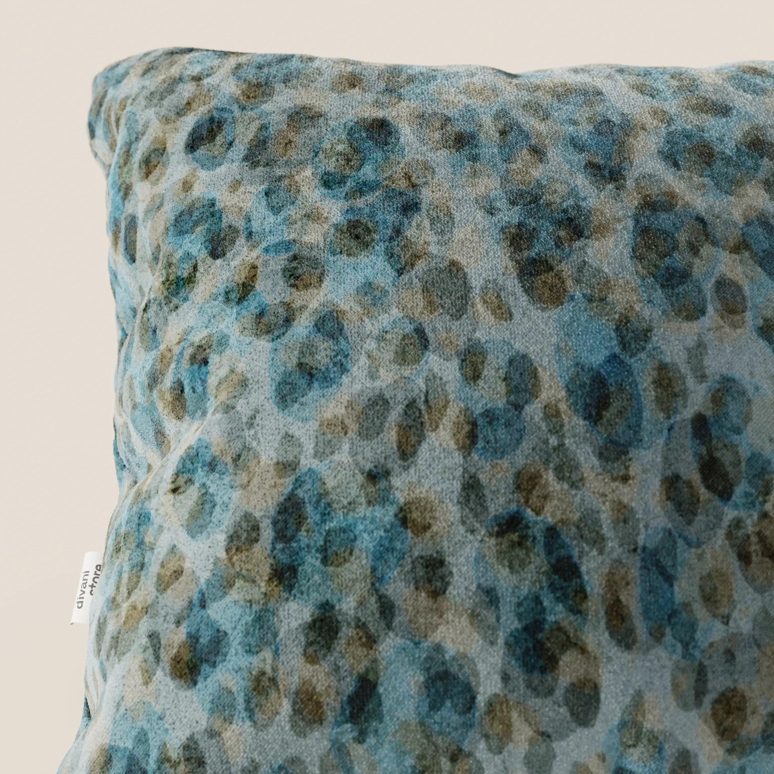 Primo piano del cuscino Brush 45x45 cm di Divani.Store in morbido velluto blu impermeabile, caratterizzato da un motivo maculato acquerellato e da una piccola etichetta bianca con testo nero parzialmente visibile sul bordo.