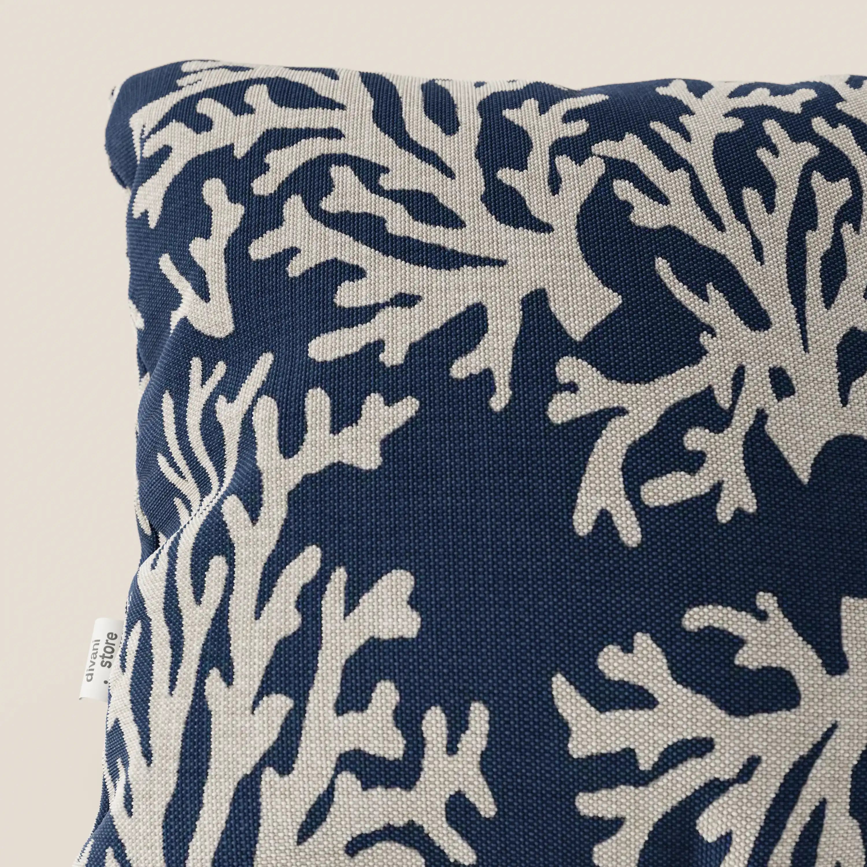 Primo piano di un cuscino Divani.Store Nautilus 45x45 in tessuto impermeabile coralli, caratterizzato da uno sfondo blu navy con un motivo di barriera corallina bianca e un'etichetta bianca visibile sul bordo sinistro.
