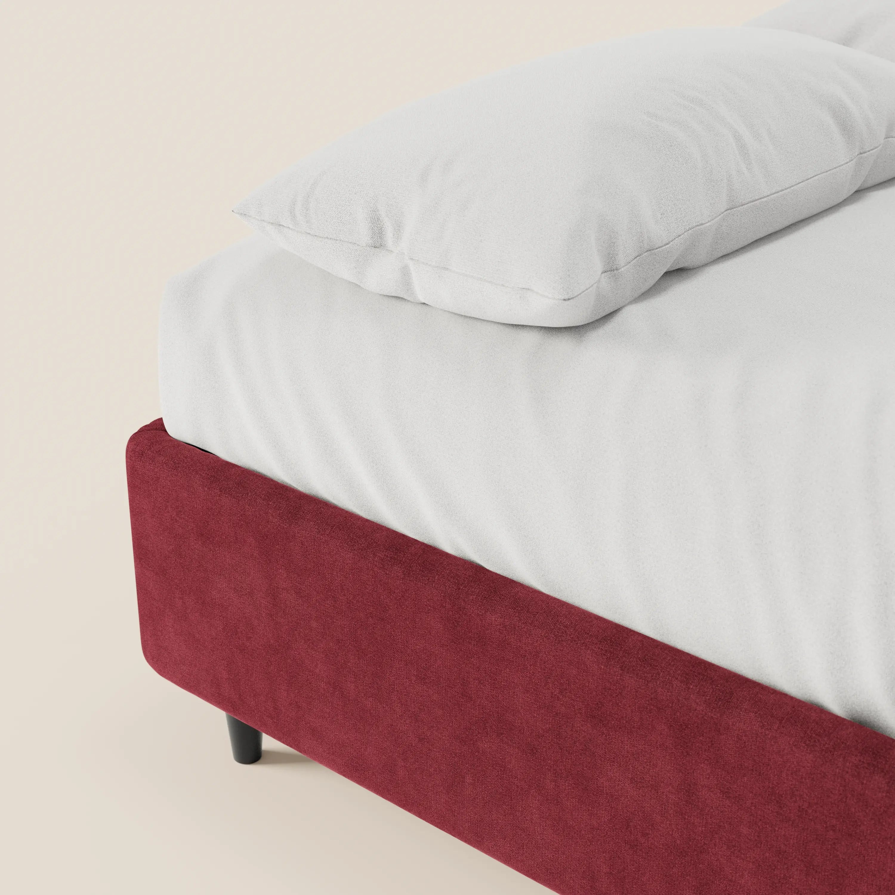 Un primo piano del letto sommier imbottito ERON con contenitore di Letti.store mostra un morbido cuscino bianco e un lenzuolo con angoli sulla sua base in tessuto impermeabile certificato Oeko-Tex di un rosso intenso, con piedini neri corti: l’ideale per chi cerca un letto elegante e senza testiera.