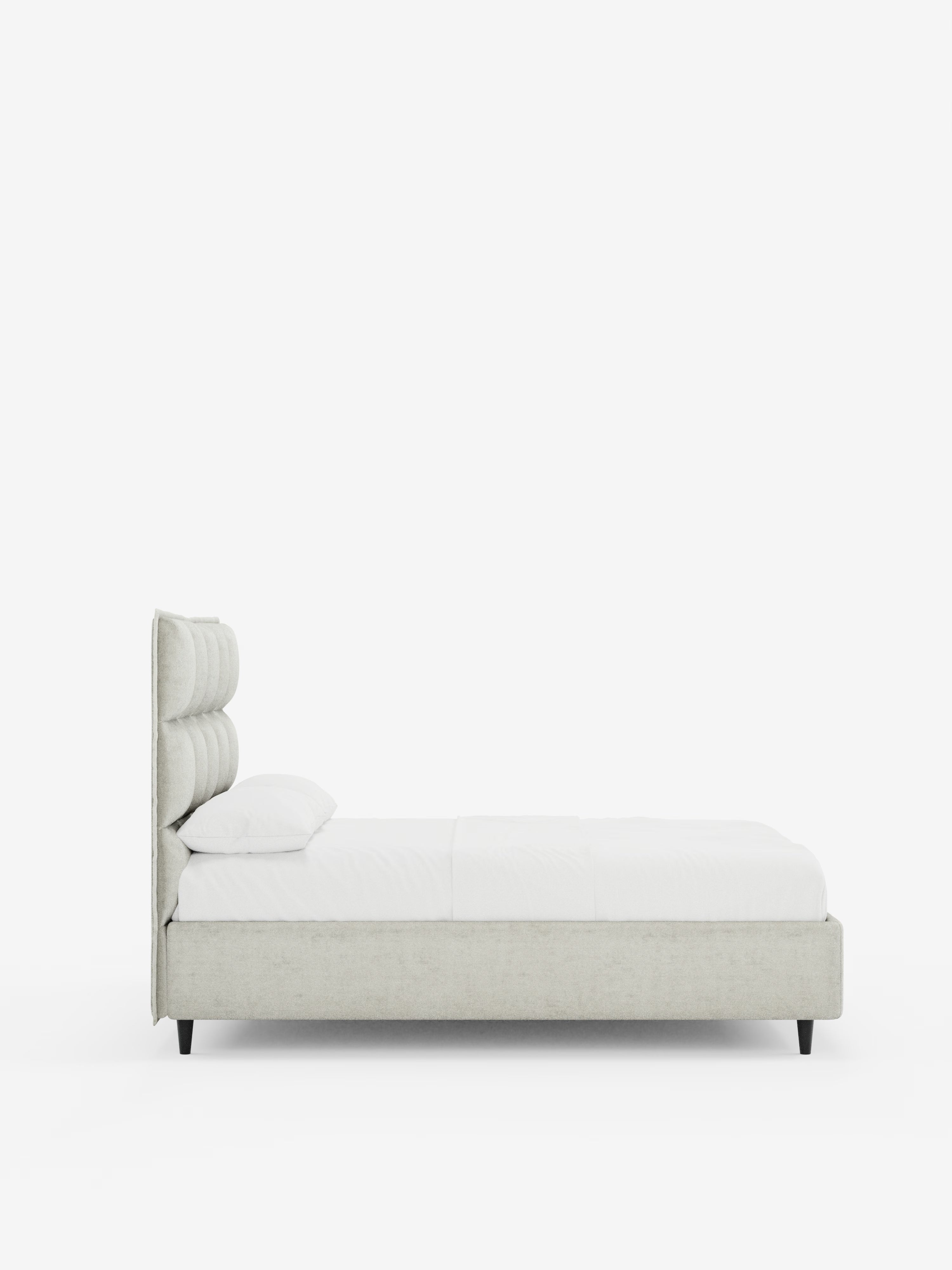 Vista laterale del letto imbottito AURO con contenitore e testiera alta capitonné in ciniglia Oeko-Tex® di Letti.store, caratterizzato da una moderna testiera alta imbottita di colore grigio chiaro con effetto capitonné, rivestita in tessuto ciniglia. Il letto è abbinato a biancheria da letto bianca, un unico cuscino e gambe basse in legno. Sullo sfondo bianco a tinta unita, spicca il design minimalista del letto.