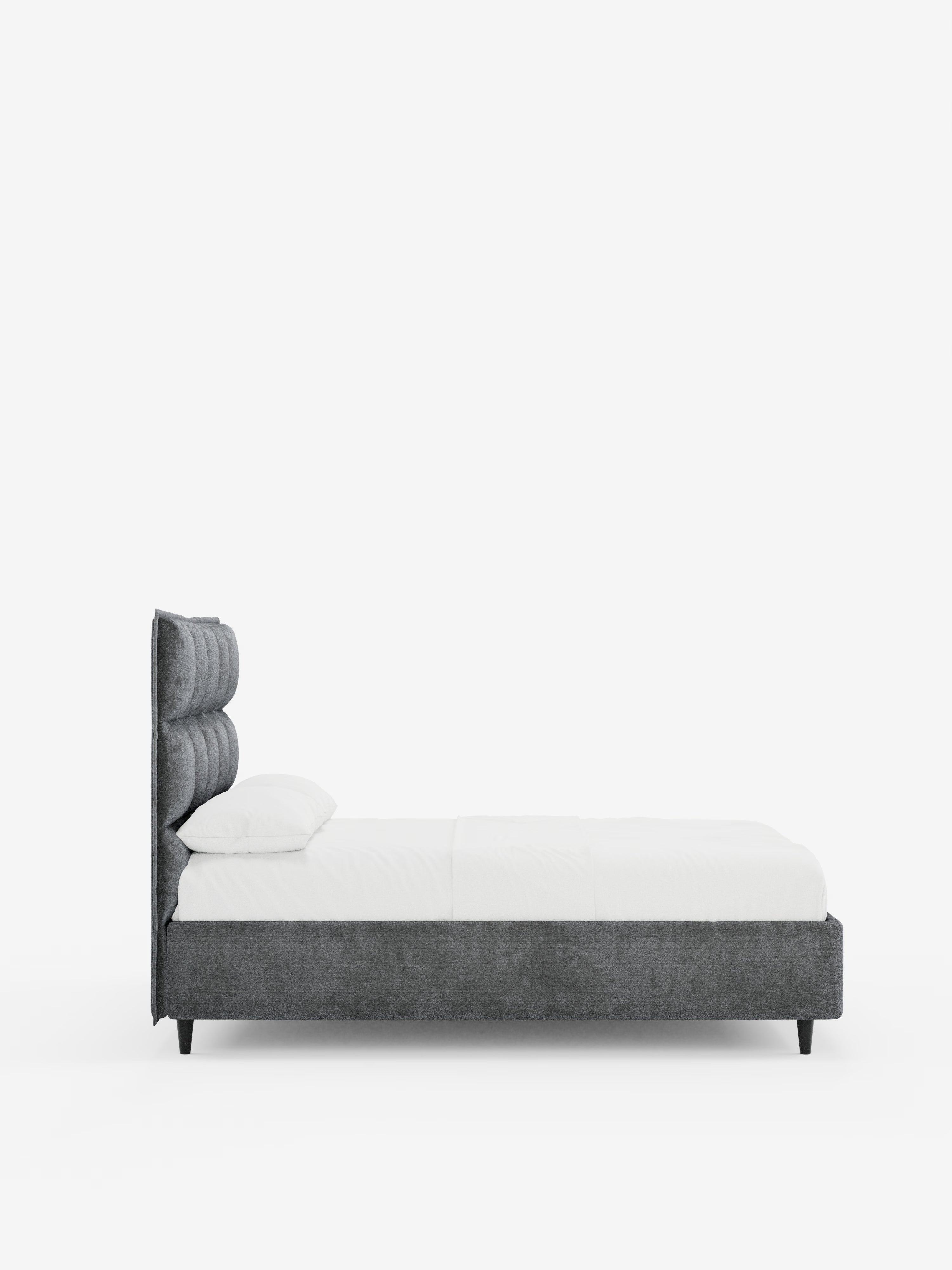 Una vista di profilo del letto imbottito AURO con contenitore e testiera alta capitonné in ciniglia Oeko-Tex® di Letti.Il modello, presentato nel negozio, presenta una testiera e una struttura imbottite in grigio scuro con un caratteristico design capitonné alto, biancheria da letto e cuscini bianchi, posizionato a terra su corte gambe nere su uno sfondo bianco a tinta unita.