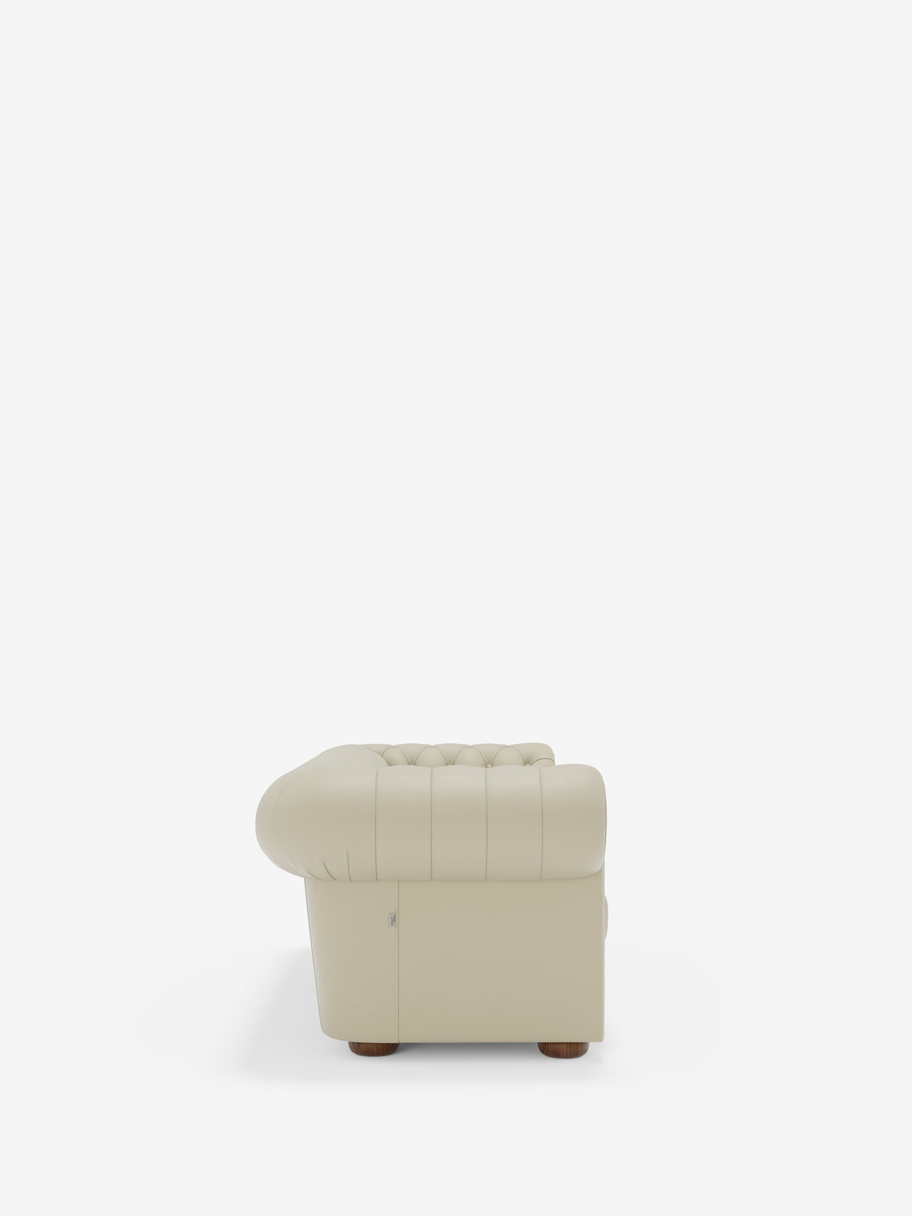 Vista laterale del Chester – Divano Chesterfield in vera pelle spessorata a 2 e 3 posti di Divani.Store, caratterizzato da rivestimento in vera pelle trapuntata color beige chiaro, braccioli arrotondati e piedini bassi in legno scuro. La poltrona presenta un design morbido e imbottito nel classico stile Chesterfield ed è posizionata al centro su uno sfondo bianco per mettere in risalto il suo elegante profilo laterale.