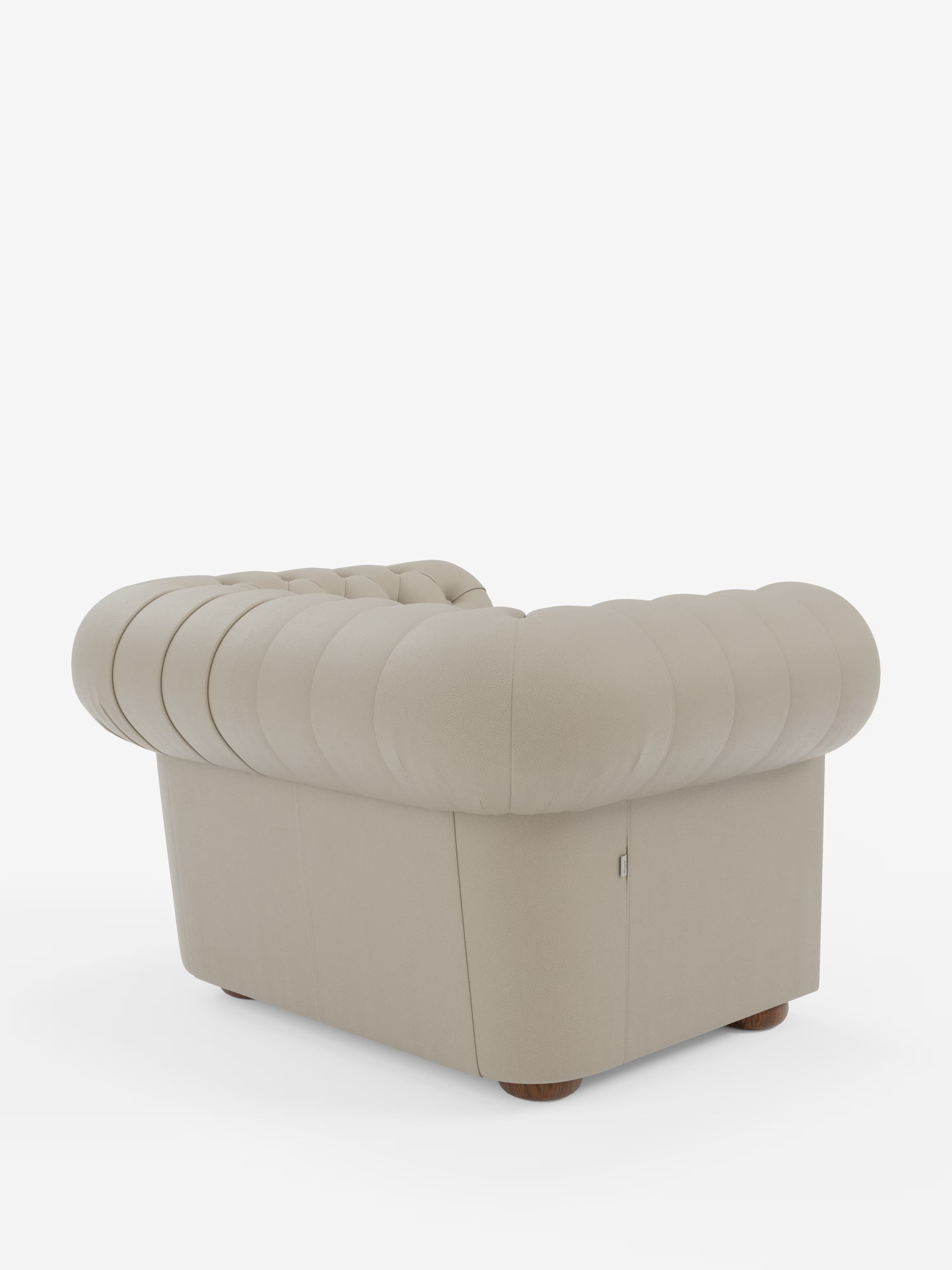 Una poltrona Chester beige in vera pelle spessorata con capitonné artigianale di Divani.Store è ripresa di spalle da un'angolazione obliqua. La poltrona presenta uno schienale arrotondato e imbottito, braccioli con discrete cuciture verticali e gambe in legno, ed è posizionata su uno sfondo bianco.
