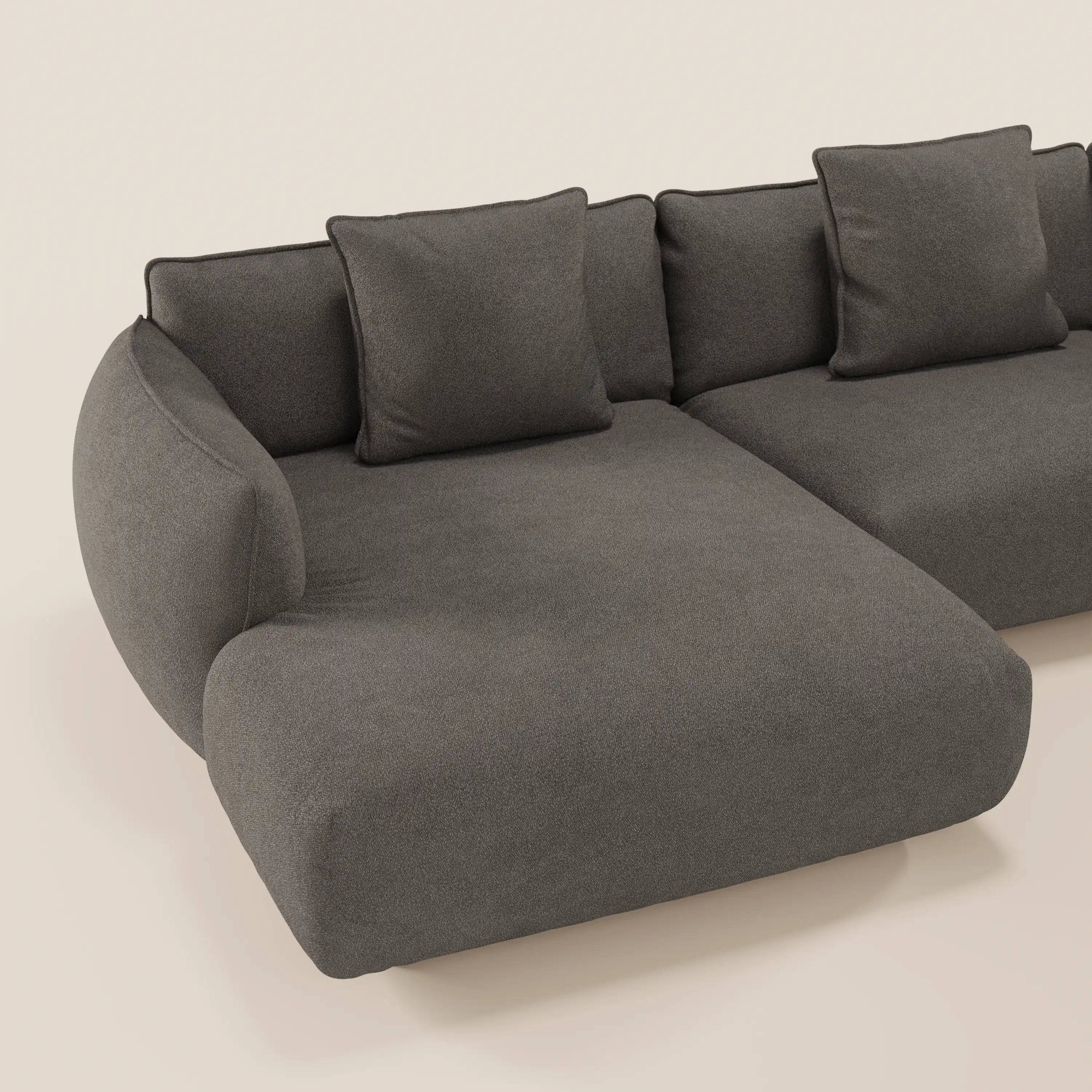 Il divano Nimbus angolare panoramico di Divani.Store è un moderno divano modulare grigio scuro con chaise longue, bracciolo curvo, tessuto antimacchia (t33) e tre cuscini di schienale, su uno sfondo beige chiaro. Prodotto in Italia.