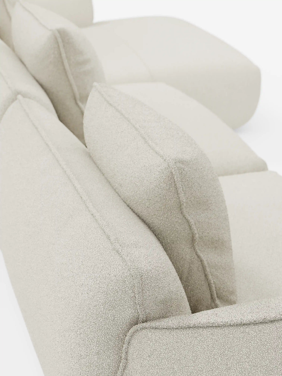 Primo piano del divano angolare con penisola componibile Nimbus di Divani.Store, rivestito in un morbido tessuto T33 effetto cotone, resistente alle macchie, in beige chiaro. I morbidi cuscini sono leggermente inclinati, mettendo in risalto la texture morbida del divano e le sue linee pulite e minimaliste. Lo sfondo è bianco brillante, a tinta unita.