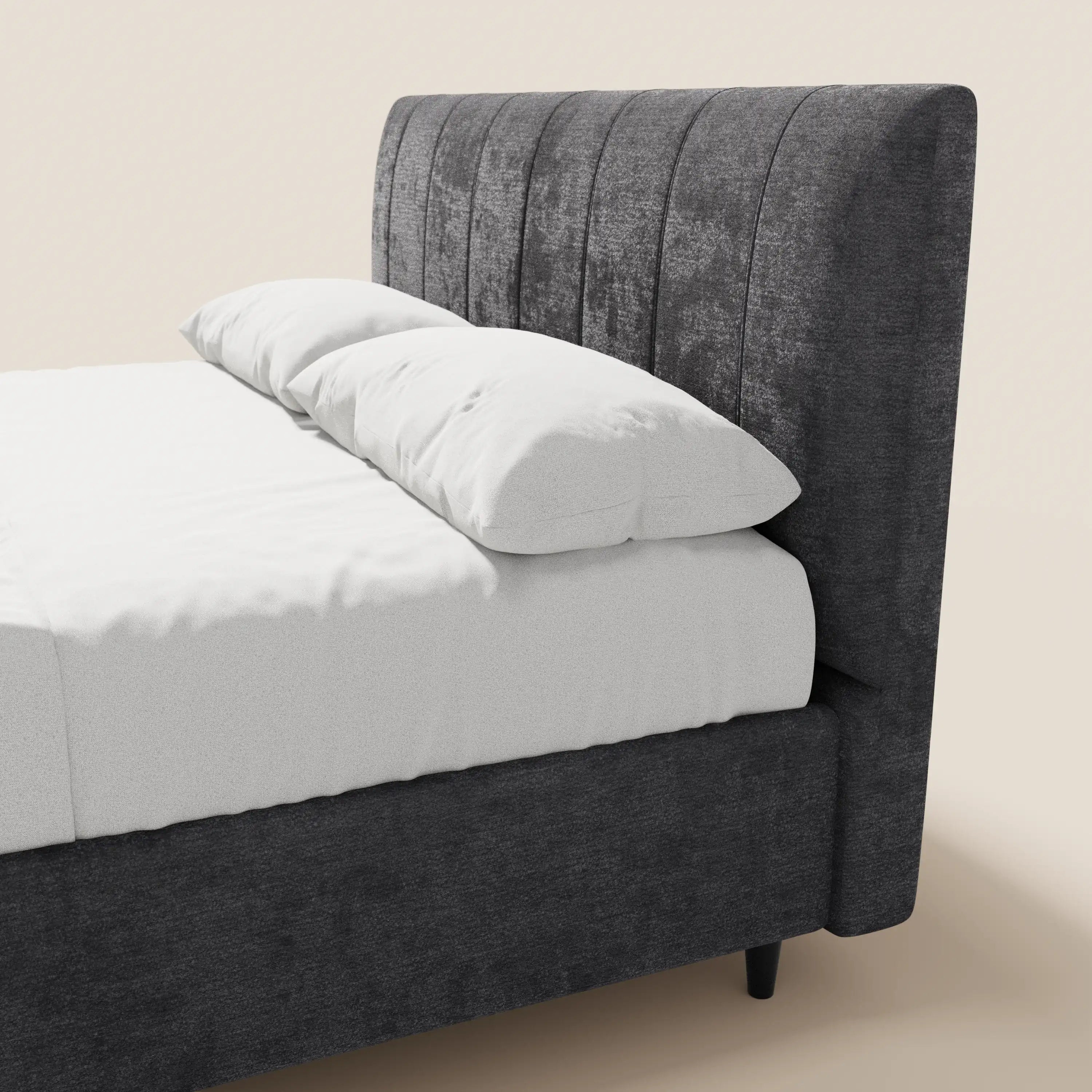Il letto imbottito ONYX di Letti.store presenta una testiera grigio scuro certificata Oeko-Tex® con canalature verticali, una base contenitore, un lenzuolo bianco e due cuscini sovrapposti su uno sfondo chiaro e semplice. È visibile solo il suo elegante lato sinistro.
