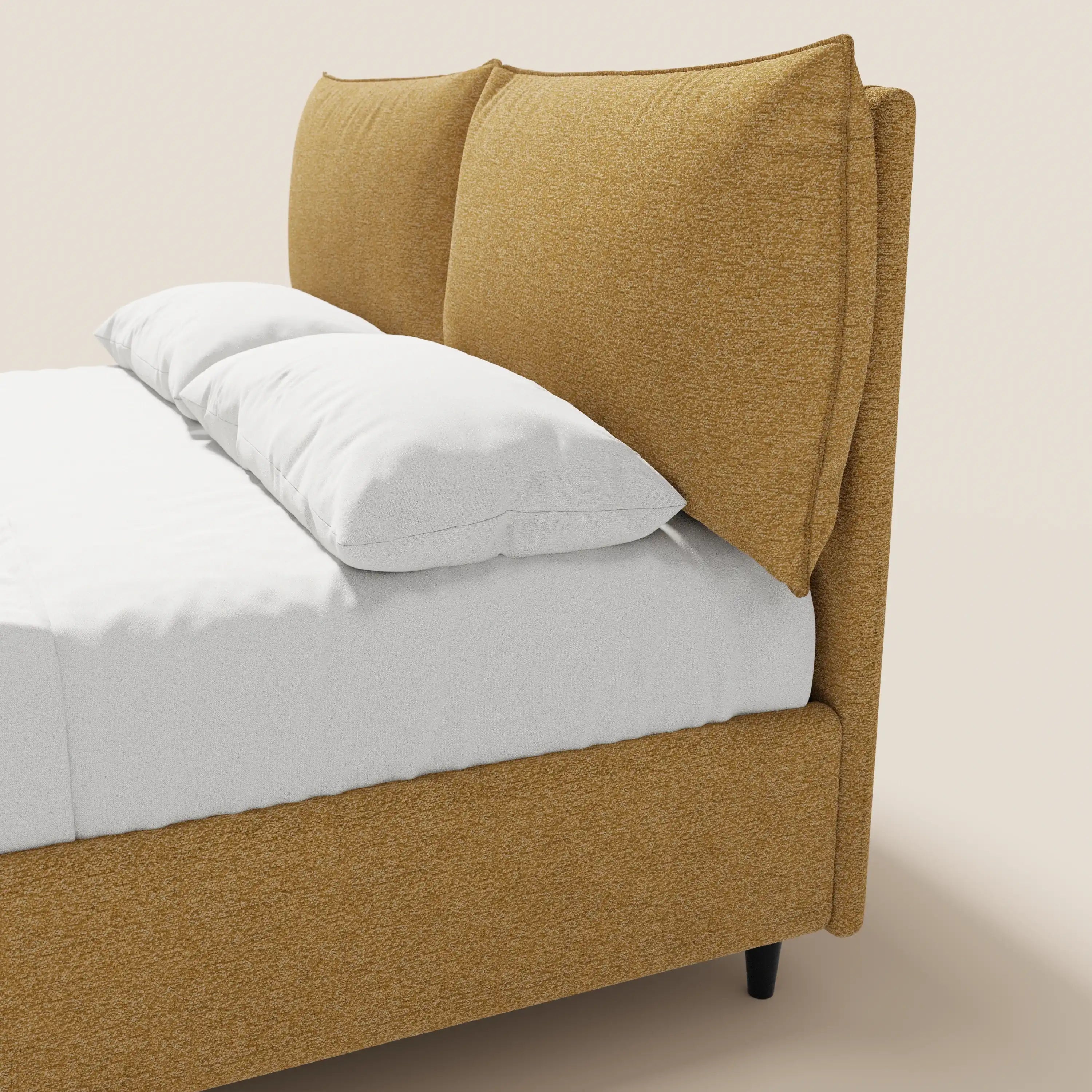 Una vista laterale ravvicinata del letto imbottito ZAYRON di Letti.store, caratterizzato da una moderna struttura imbottita in giallo senape certificata Oeko-Tex®, da una testiera regolabile simile a un cuscino e da un vano contenitore sotto il letto, presentato con lenzuola bianche su uno sfondo beige.