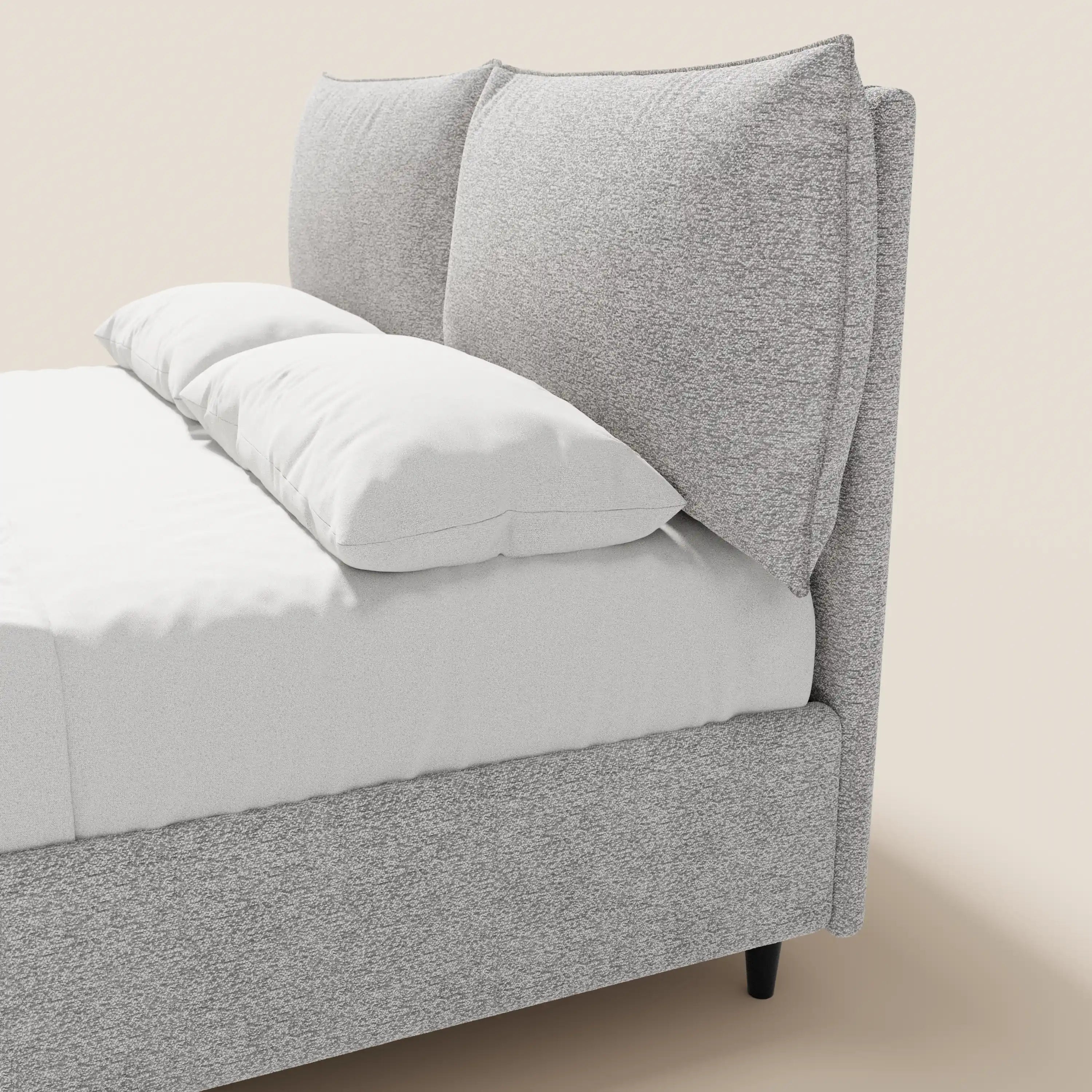 Un primo piano del letto imbottito ZAYRON di Letti.store, caratterizzato da una struttura in tessuto grigio chiaro certificato Oeko-Tex® e da morbidi cuscini regolabili. Questo letto dal design moderno è abbinato a lenzuola bianchissime che risaltano su uno sfondo beige tenue.