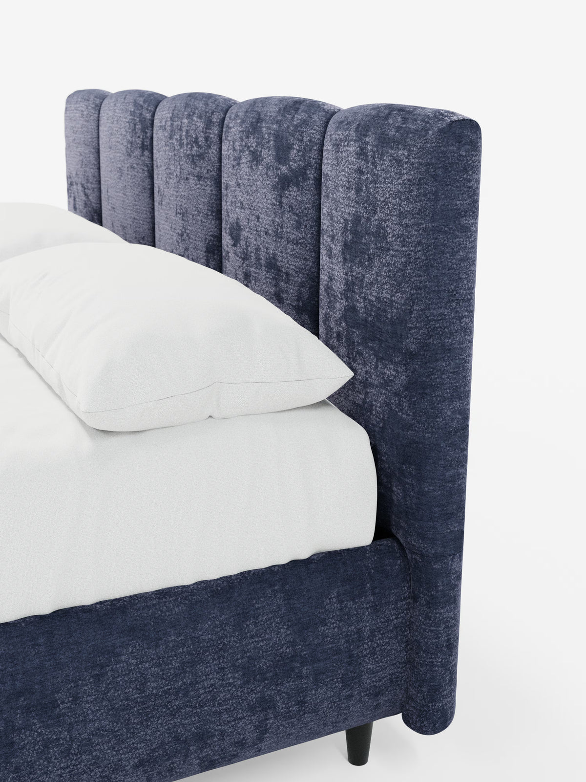 Primo piano di un angolo del letto che mette in risalto il modello ARKAM di Letti.store: un letto imbottito con vano contenitore e testiera a lavorazione verticale. Questa testiera alta e morbida, in blu navy, presenta cuciture a canalina verticali e incarna lo stile shabby chic grazie al tessuto certificato Oeko-Tex®. Il letto, rivestito con biancheria da letto bianca e con il materasso a vista, risalta sullo sfondo bianco liscio, mettendo in risalto le ricche trame e il colore intenso di questo elegante