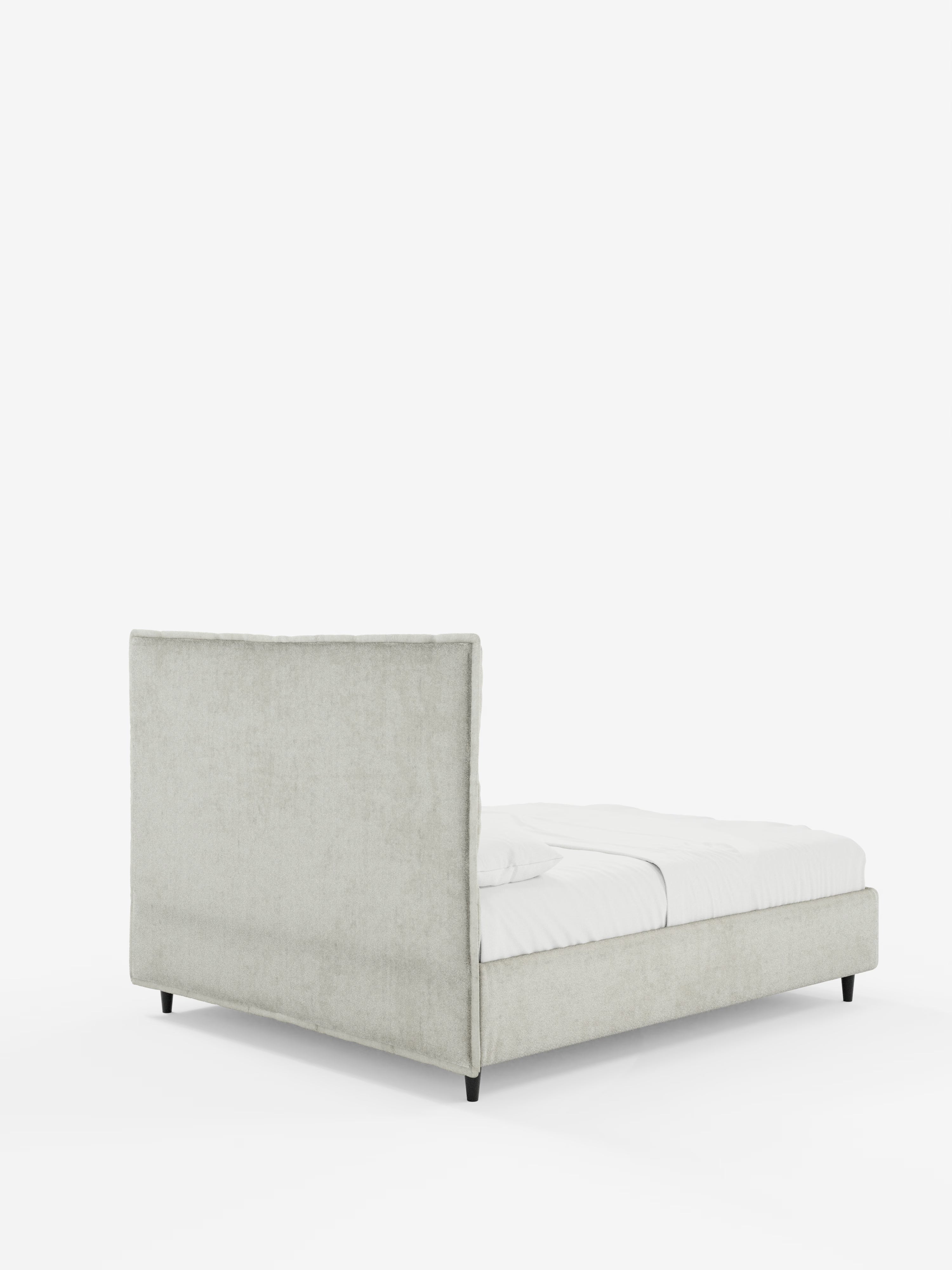 Il letto imbottito AURO con contenitore e testiera capitonné alta in ciniglia Oeko-Tex® di Letti.store è ripreso da un angolo posteriore. Questo letto a piattaforma imbottito di colore grigio chiaro presenta una testiera alta, morbidamente imbottita e capitonné, e una struttura bassa. Il materasso è rivestito con un lenzuolo con angoli bianco e una coperta, e il letto poggia su gambe corte e scure su uno sfondo bianco a tinta unita.