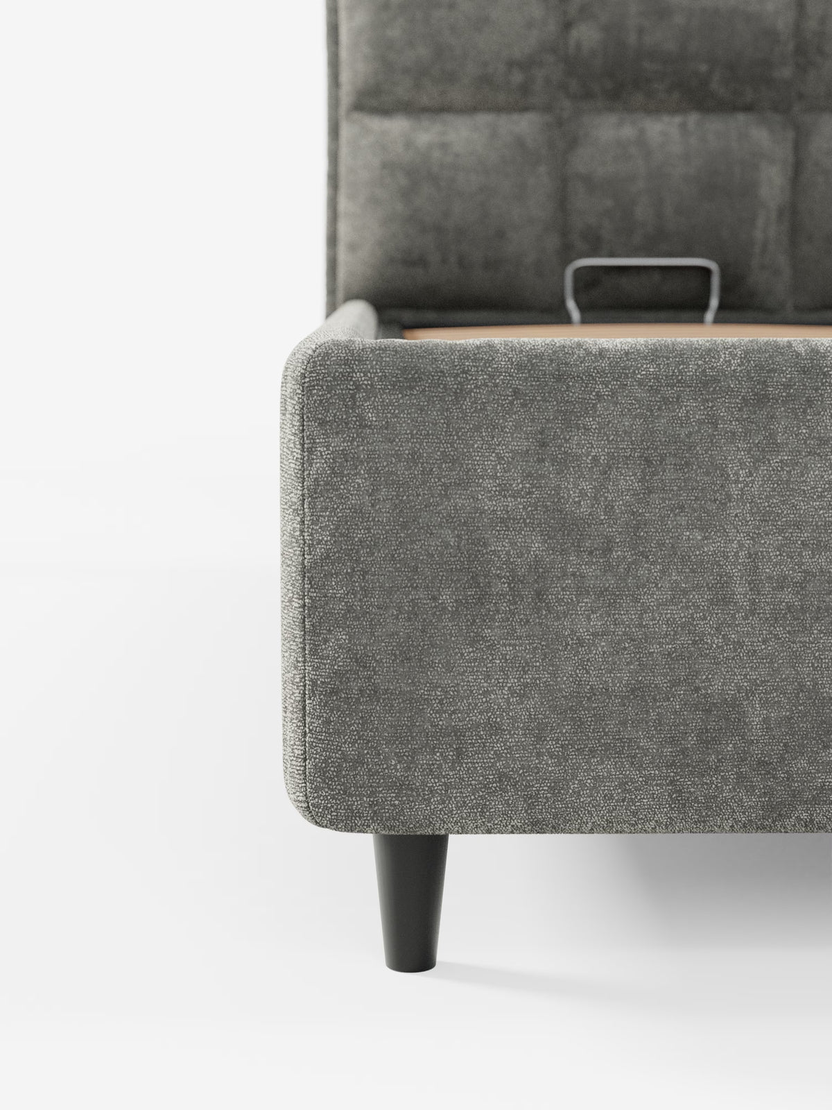 Primo piano di un letto imbottito AURO di Letti.store, caratterizzato da un angolo della struttura del letto rivestito in ciniglia grigia certificata Oeko-Tex® con tessuto testurizzato e da una gamba cilindrica in legno nero. È visibile parte della testiera alta e capitonné. Una piccola maniglia in metallo vicino al centro indica la presenza di un pratico vano contenitore per una maggiore praticità di stoccaggio. Lo sfondo è bianco a tinta unita.