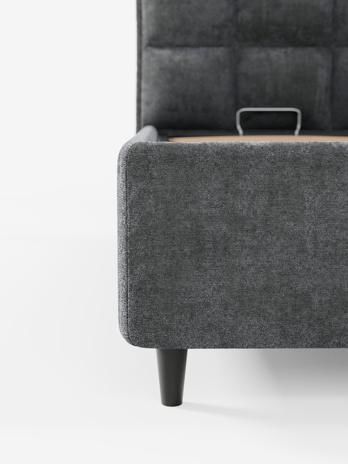 Primo piano del letto imbottito AURO con vano contenitore e testiera capitonné alta in ciniglia Oeko-Tex® di Letti.store, qui raffigurato con rivestimento in grigio moderno. Su uno sfondo bianco a tinta unita sono visibili solo l’angolo anteriore inferiore e una gamba nera affusolata, con una vista parziale della struttura metallica del vano contenitore all’interno del letto.