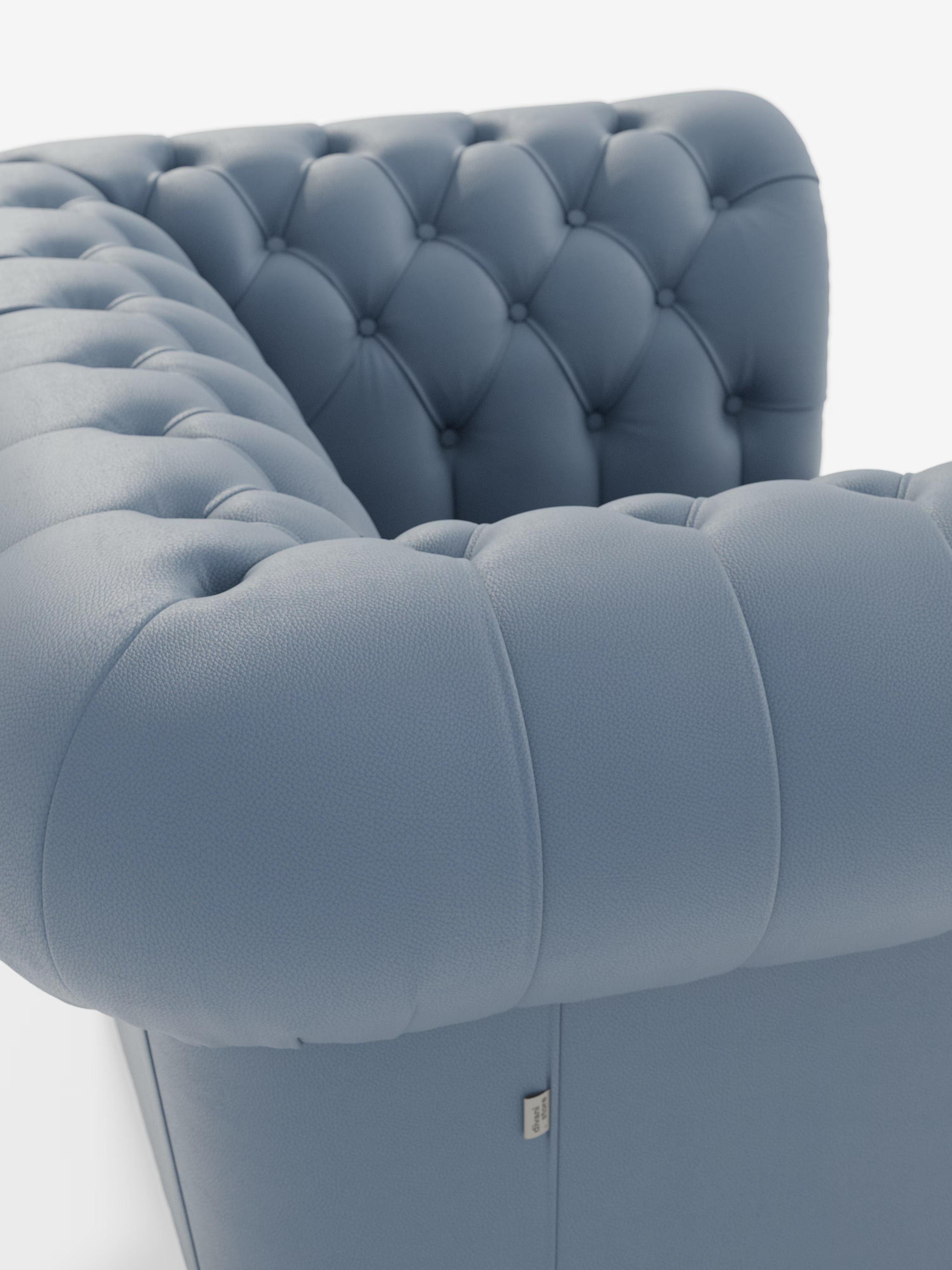 Primo piano della poltrona Chester in vera pelle spessorata con capitonné artigianale di Divani.Store: una poltrona in pelle blu trapuntata, caratterizzata da profondi bottoni realizzati a mano sullo schienale e sui braccioli. L’immagine mette in risalto la superficie liscia e ruvida al tatto e le raffinate cuciture del rivestimento che caratterizzano questo lussuoso complemento d’arredo; sul lato è apposta una piccola etichetta quadrata in tessuto.