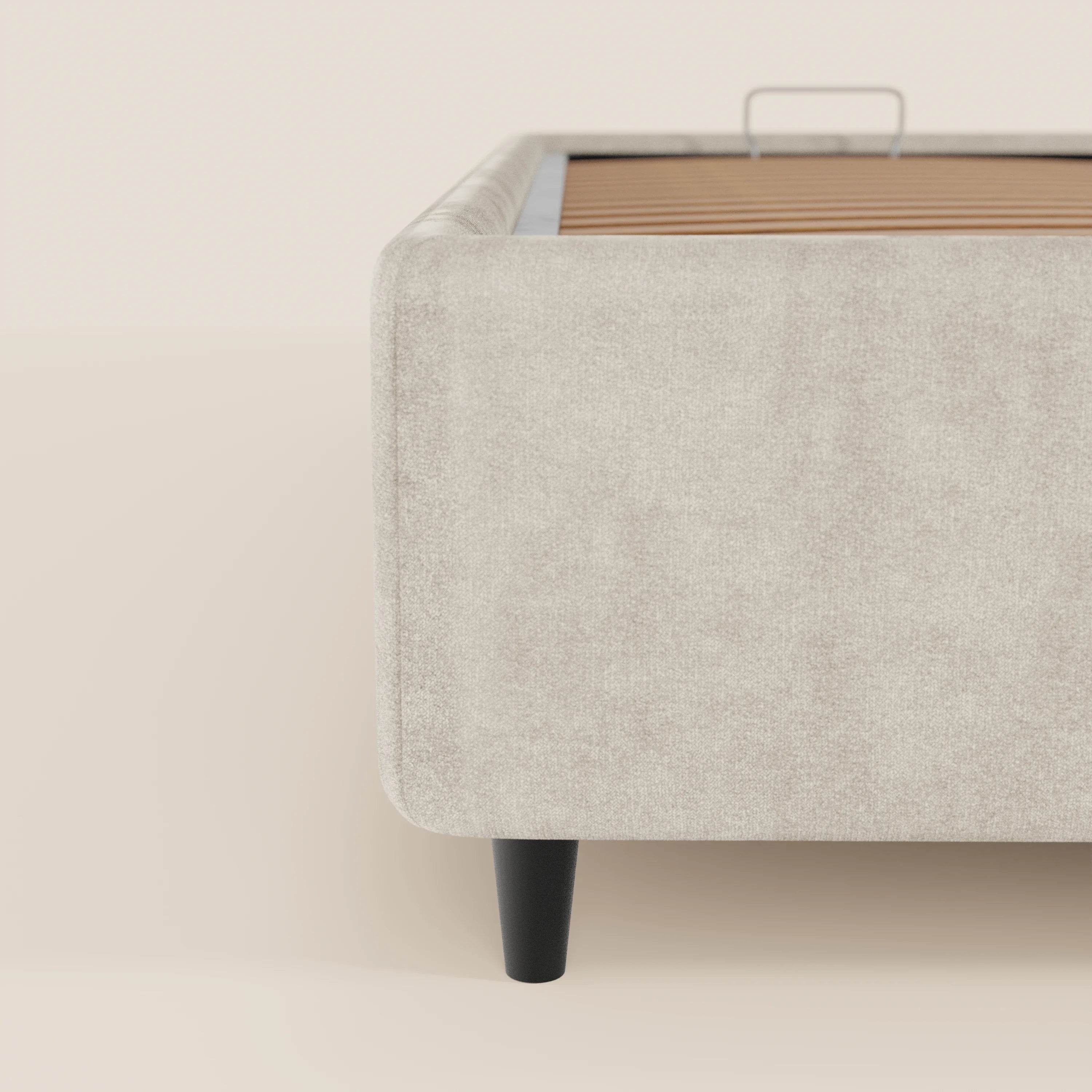 Primo piano del letto sommier imbottito ERON di Letti.store, caratterizzato da angoli arrotondati, gambe corte e angolate di colore nero e doghe in legno a vista. Rivestito in tessuto impermeabile certificato Oeko-Tex, con vano contenitore. Sfondo beige chiaro.