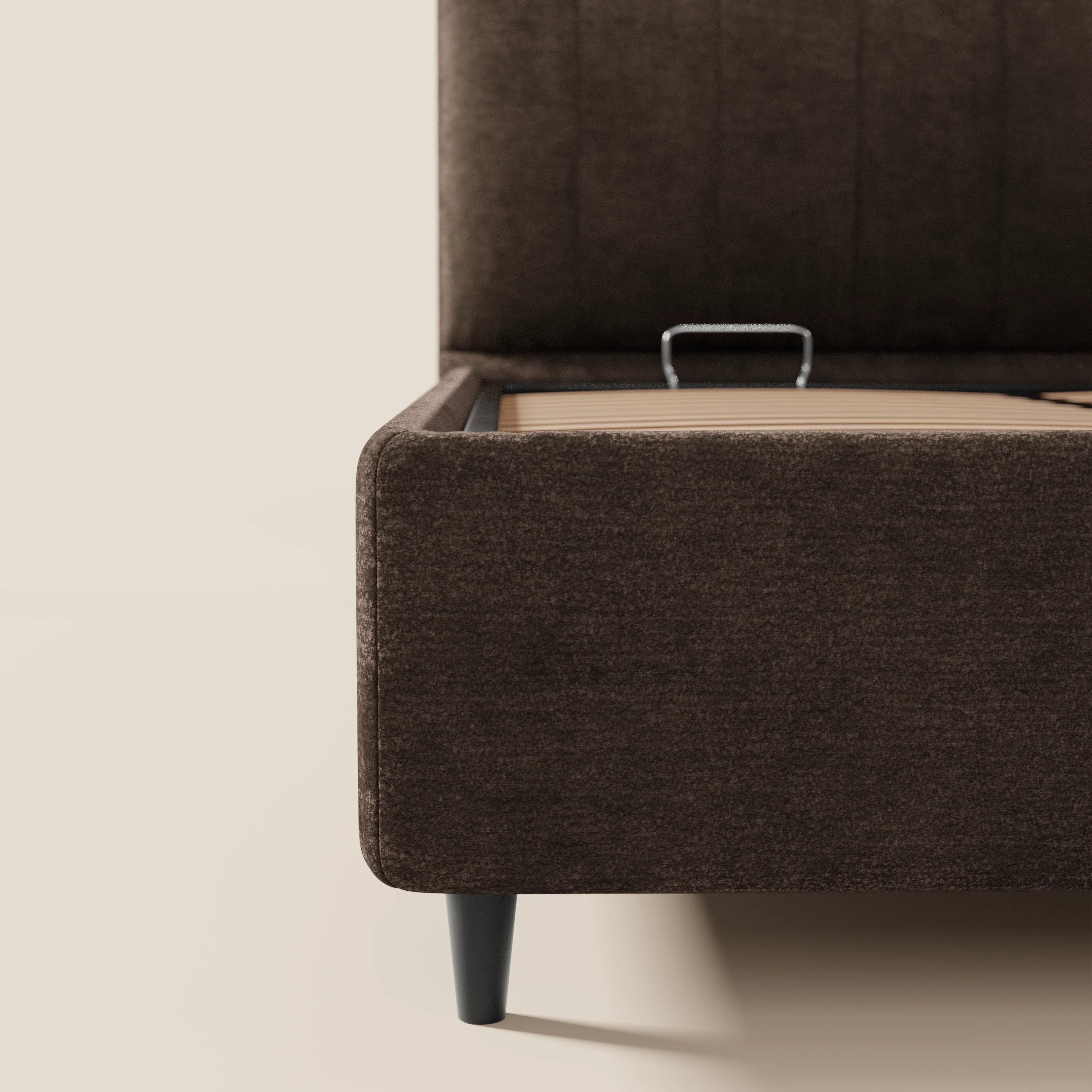 Primo piano del letto imbottito con vano contenitore ONYX di Letti.store, caratterizzato da un tessuto marrone scuro certificato Oeko-Tex®, una testiera verticale alta e imbottita, gambe affusolate nere e un vano contenitore aperto con maniglia. Sfondo di pareti e pavimento in tinta unita bianco sporco.