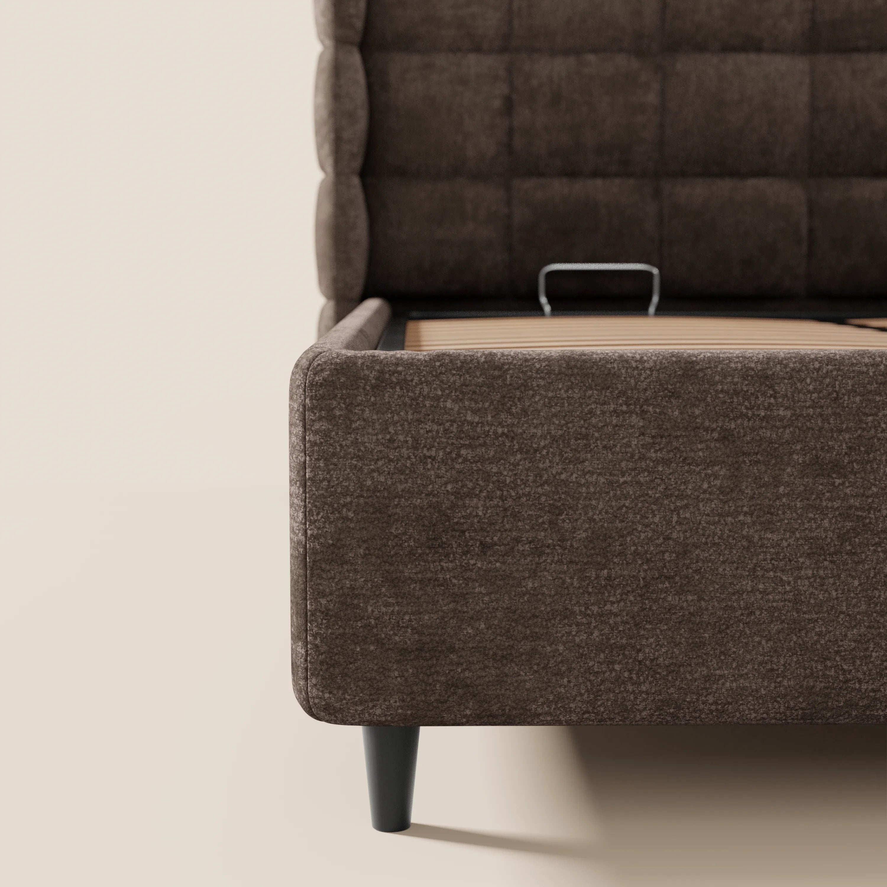 Un primo piano del letto imbottito RAGNAR con contenitore e testiera capitonné avvolgente di Letti.store, realizzato in tessuto marrone scuro certificato Oeko-Tex®, caratterizzato da quadrati trapuntati e gambe nere affusolate, ne mette in risalto l'elegante struttura e la trama.