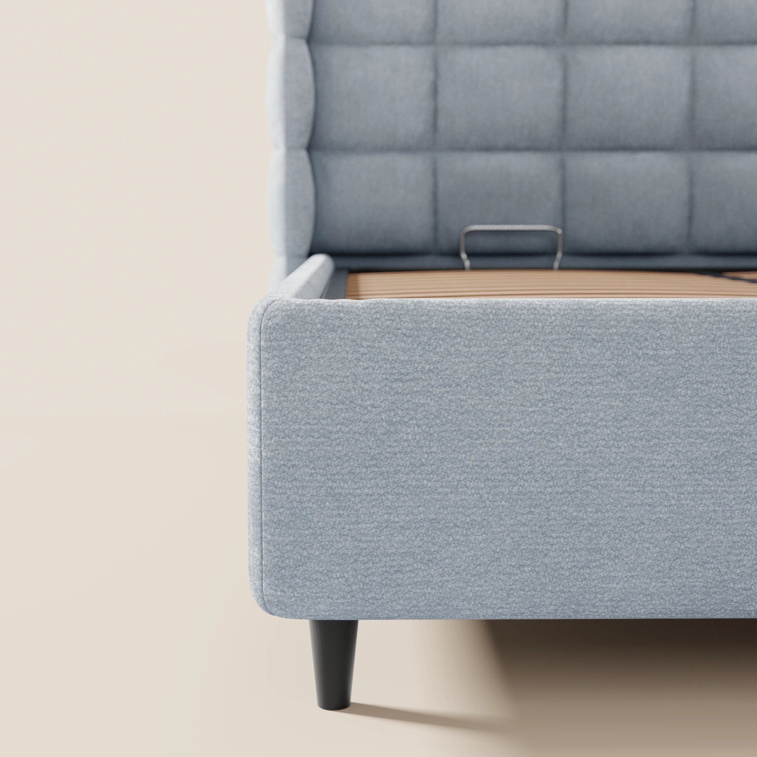 Primo piano del letto imbottito RAGNAR con contenitore e testiera capitonné avvolgente di Letti.store, realizzato in tessuto azzurro certificato Oeko-Tex®, che mostra la testiera imbottita, la sponda laterale e la gamba affusolata nera su uno sfondo beige.