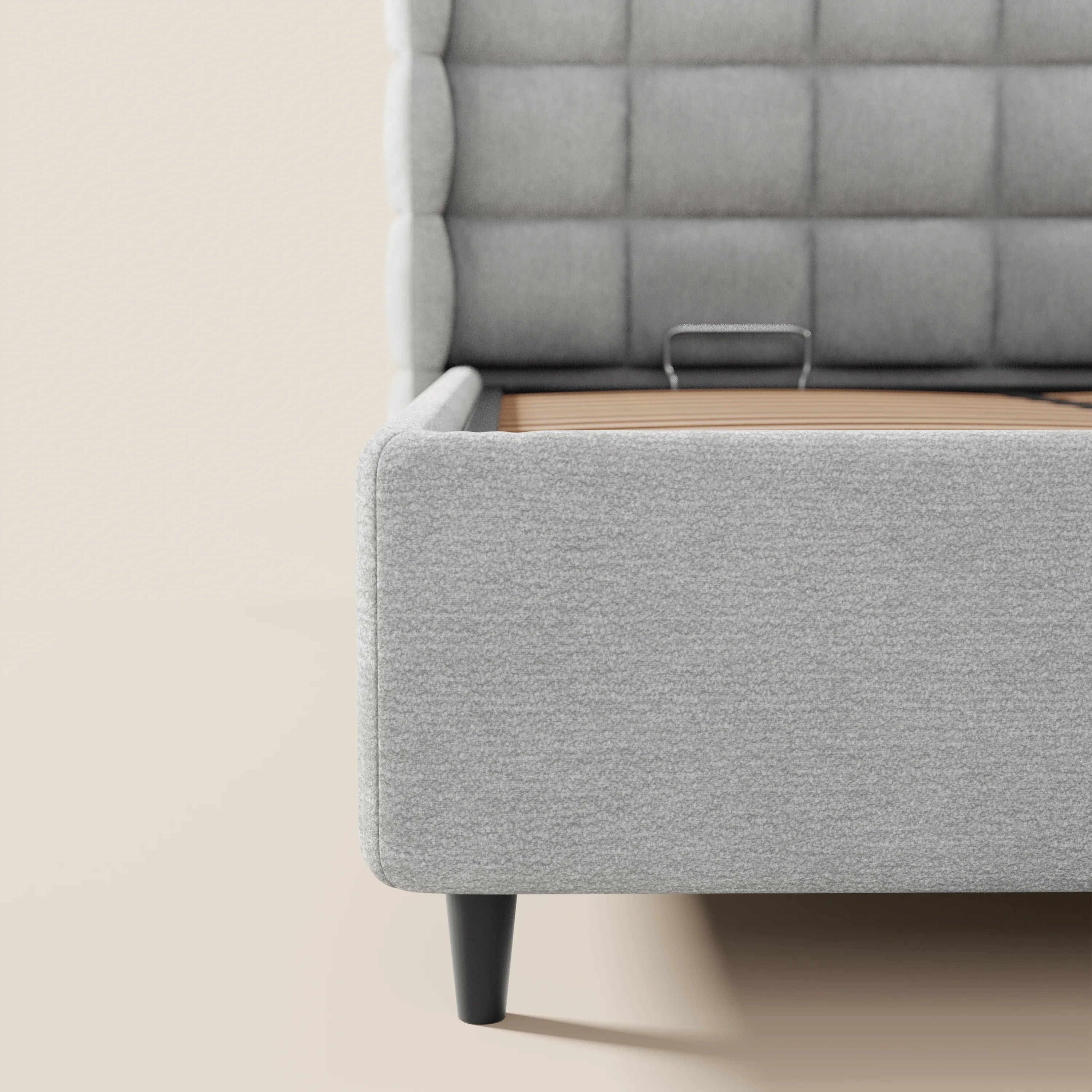 L'immagine mostra un primo piano del letto imbottito RAGNAR di Letti.store, caratterizzato da un'avvolgente testiera capitonné in elegante tessuto grigio certificato Oeko-Tex®. Su uno sfondo beige sono visibili solo la metà sinistra e le corte gambe affusolate di colore nero.