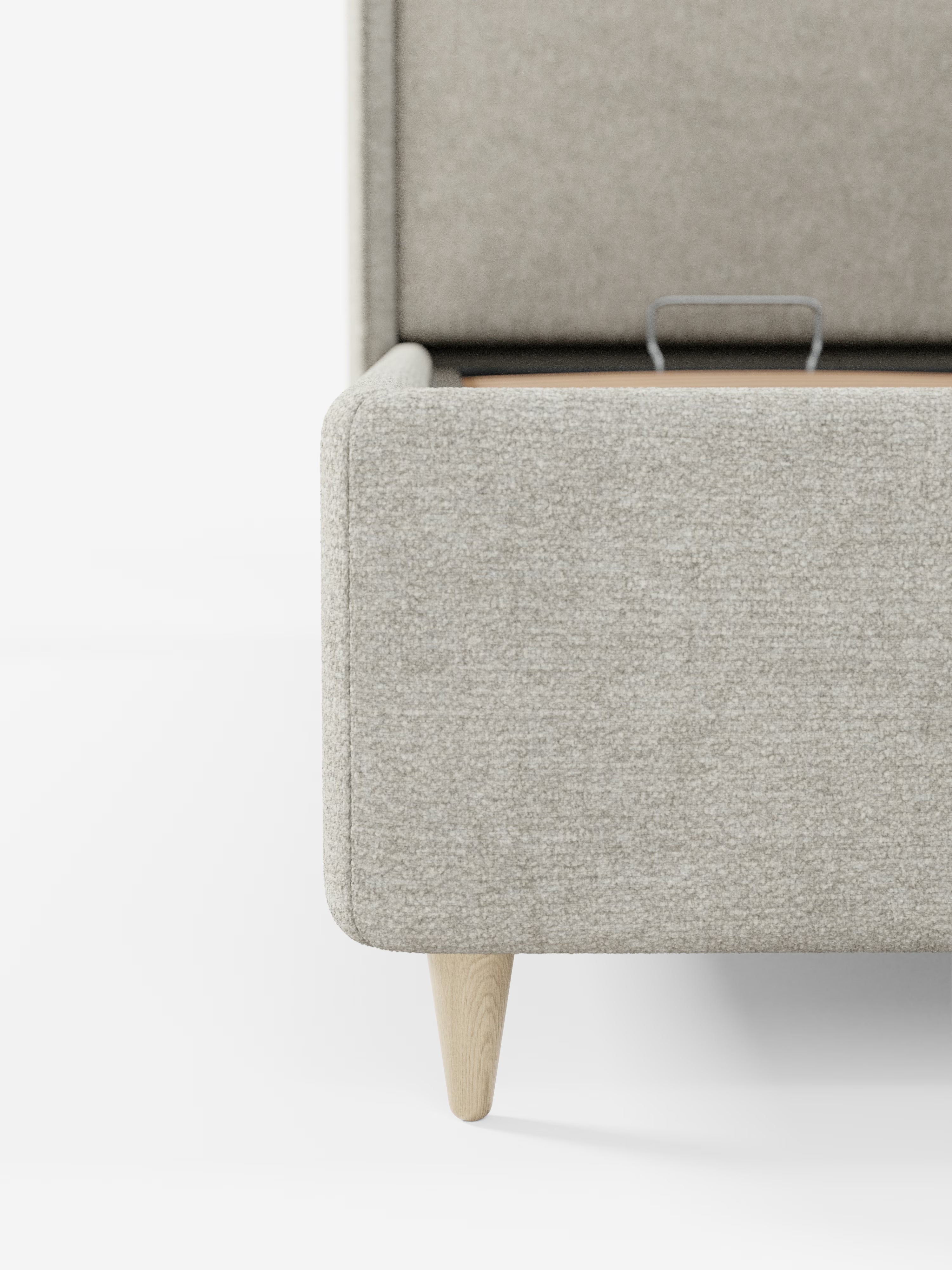 Primo piano dell'angolo del letto imbottito bicolore VALKIRIA con vano contenitore di Letti.store, caratterizzato da gambe in legno chiaro. La struttura imbottita presenta un pratico vano contenitore con il coperchio leggermente socchiuso, che lascia intravedere un'elegante maniglia in metallo su sfondo bianco.