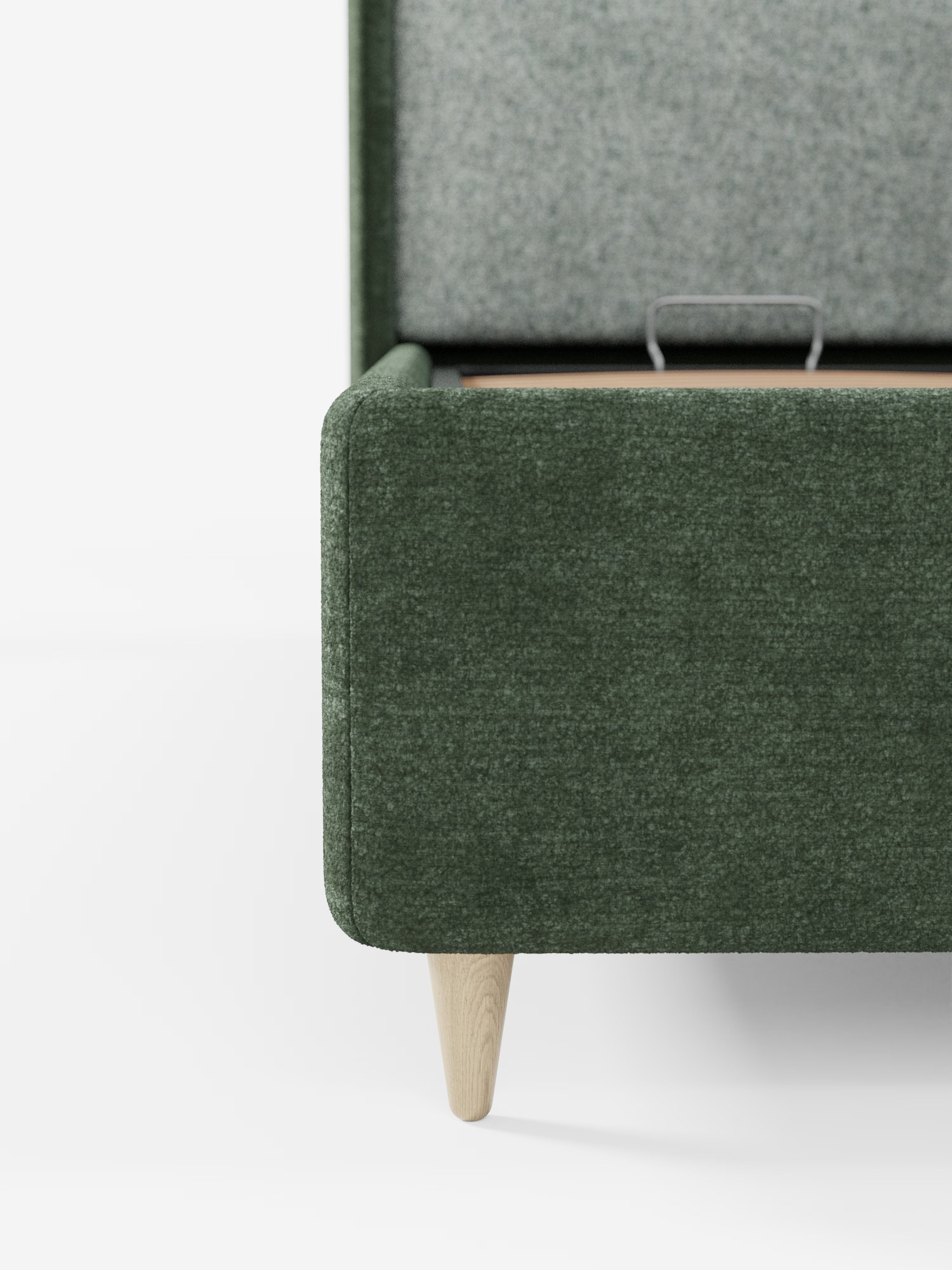 Primo piano di un angolo imbottito verde del letto VALKIRIA bicolore con vano contenitore di Letti.store, caratterizzato da una gamba affusolata in legno chiaro e parzialmente aperto per mostrare il suo spazioso vano contenitore. Il design moderno, con testiera curva avvolgente e cuscini dello schienale, risalta su uno sfondo bianco a tinta unita.