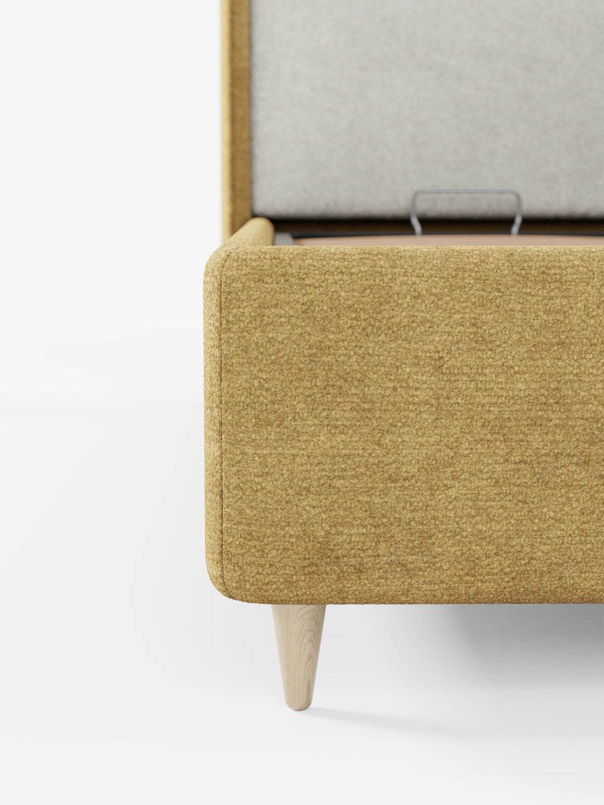 Primo piano di una panca contenitore imbottita color giallo senape con gambe in legno chiaro su sfondo bianco. Il piano è parzialmente aperto, lasciando intravedere un coperchio interno grigio chiaro e un meccanismo a cerniera in metallo simile a quello presente nel letto imbottito bicolore con vano contenitore VALKIRIA di Letti.store. L’aspetto testurizzato del tessuto riprende le raffinate finiture dei cuscini schienali e della testiera avvolgente, tipici dei modelli di questo marchio.
