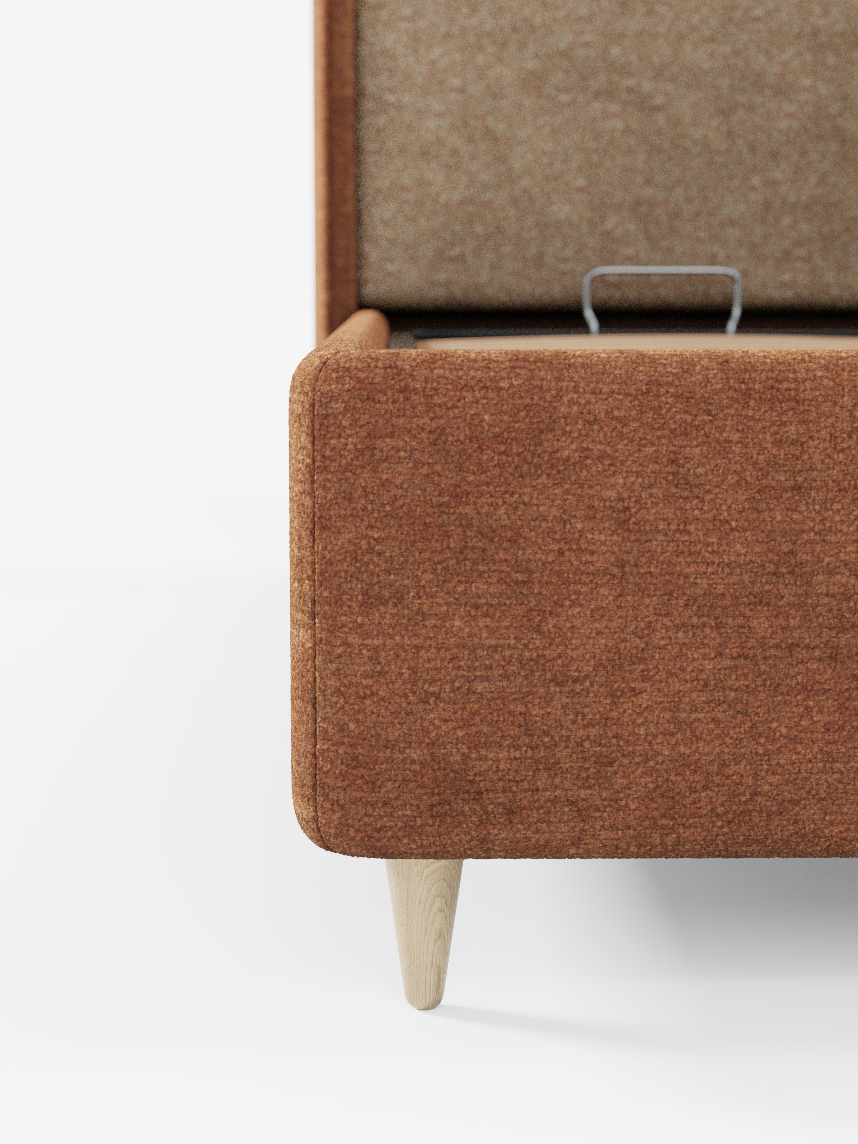 Un primo piano del letto imbottito bicolore con vano contenitore VALKIRIA di Letti.store, che mette in risalto il rivestimento color ruggine e le gambe in legno chiaro. Il coperchio è aperto e rivela un vano contenitore rivestito in tessuto con una maniglia in metallo. Su uno sfondo bianco, l’immagine mette in risalto le linee morbide della panca, che richiamano la testiera curva avvolgente e i cuscini dello schienale, evocando un elegante letto imbottito italiano con contenitore.