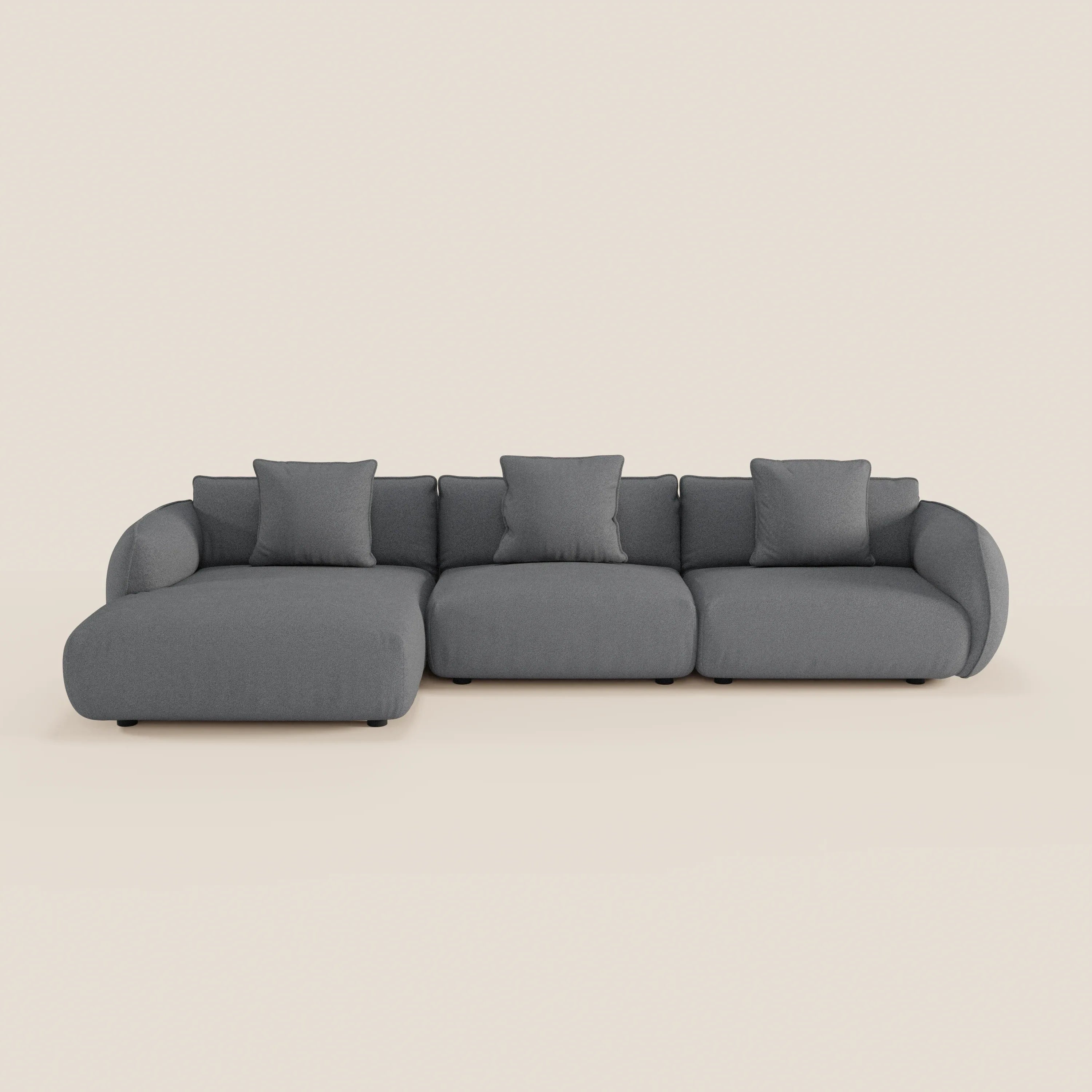 Il divano angolare Nimbus con penisola componibile in morbido tessuto effetto cotone smacchiabile T33 di Divani.Store è un moderno divano grigio scuro a tre posti, con chaise laterale sinistra, braccioli arrotondati e quattro cuscini quadrati su sfondo bianco sporco.