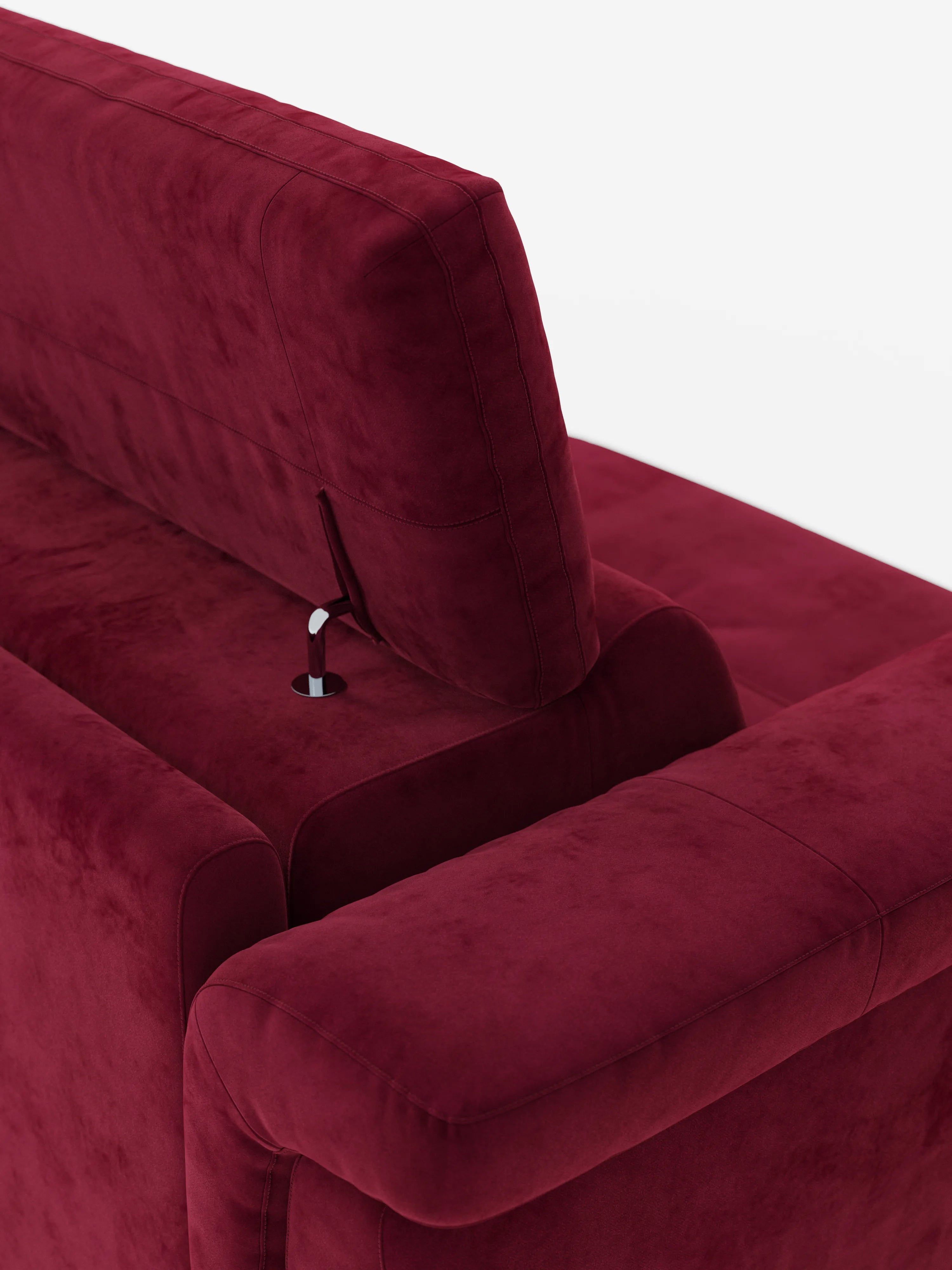 Un primo piano dello schienale e del bracciolo del divano letto di design Capri con piedini alti di Divani.Store mette in risalto il suo rivestimento in morbido tessuto effetto velluto smacchiabile di un rosso intenso (tessuto effetto velluto T30). Una leva metallica situata vicino al poggiatesta regolabile lascia intuire le sue caratteristiche ergonomiche. Su uno sfondo bianco a tinta unita, le finiture morbide e lussuose del divano e le cuciture pulite incarnano la classica maestria artigianale italiana.
