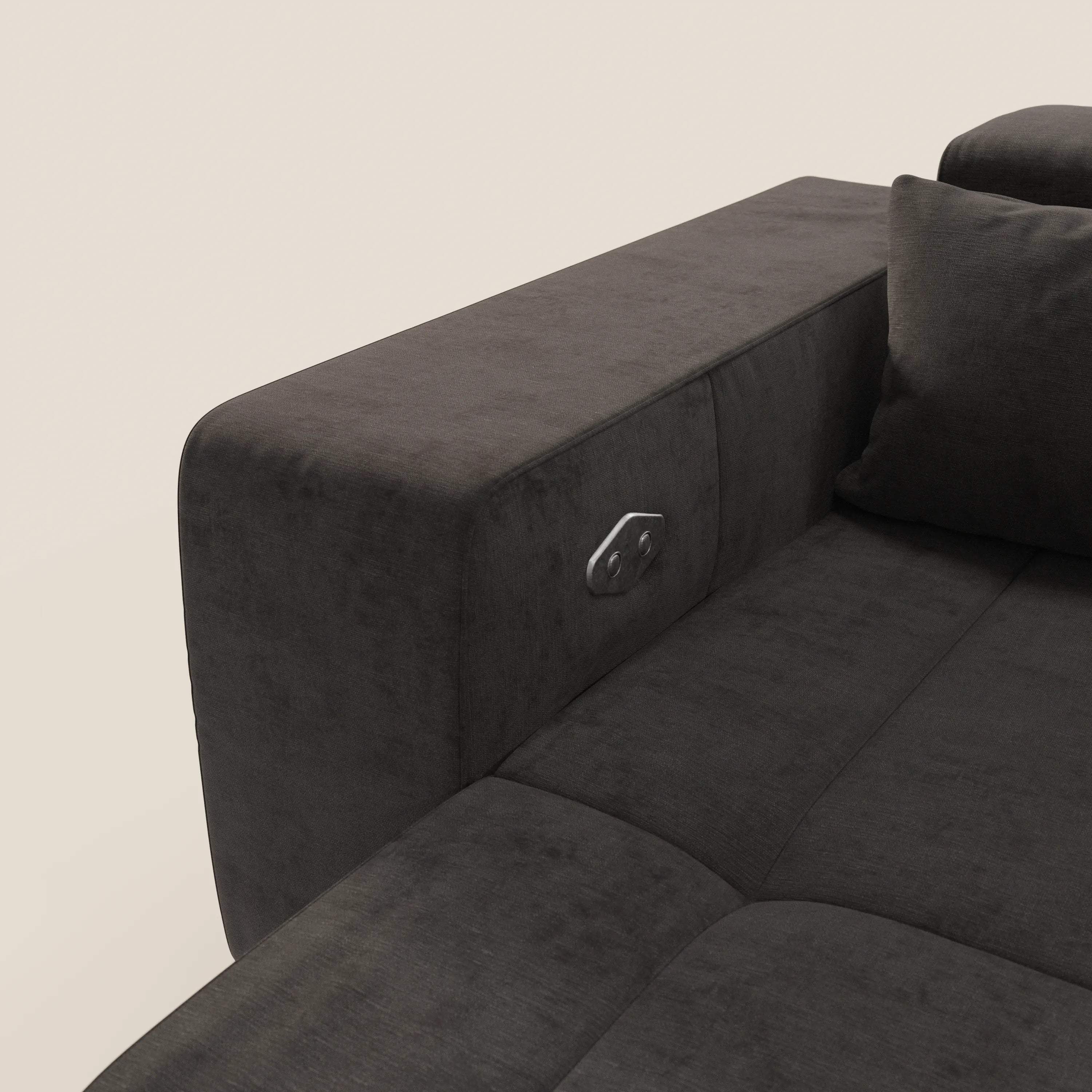 Un primo piano del Monarch Divano con Relax Elettrico di Divani.Store in grigio scuro mostra un bracciolo in peluche con un bottone metallico, parte dello schienale con cuscino, su uno sfondo bianco sporco.