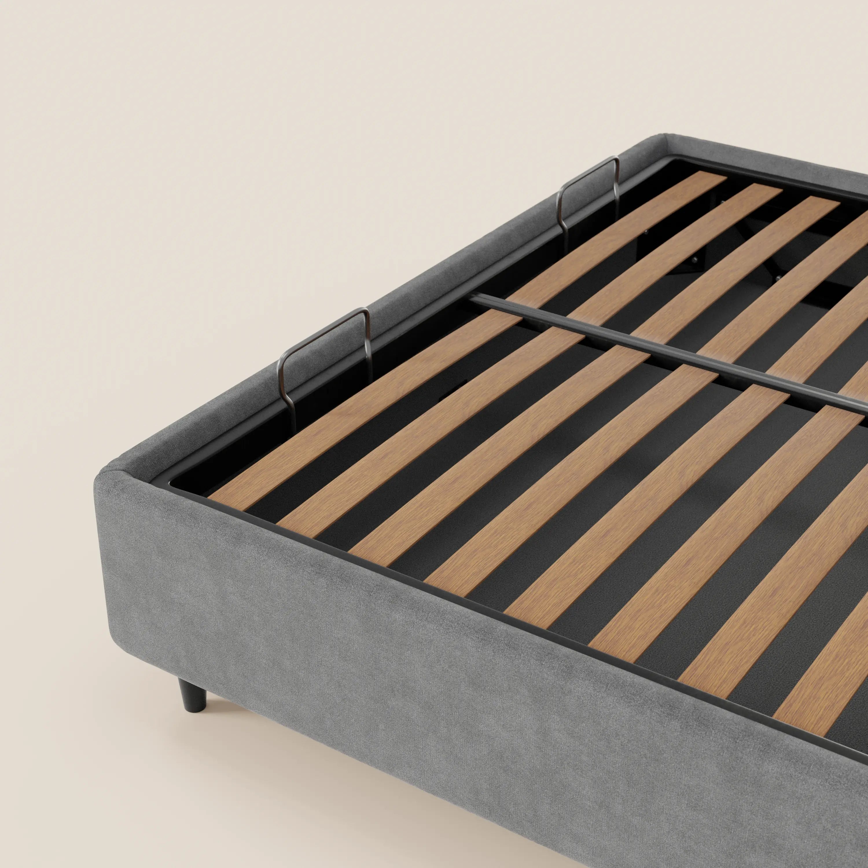 Un primo piano del letto sommier imbottito ERON di Letti.store, caratterizzato da una base in tessuto grigio impermeabile certificato Oeko-Tex, gambe nere, doghe in legno e due staffe metalliche sul telaio interno, su uno sfondo beige chiaro a tinta unita.