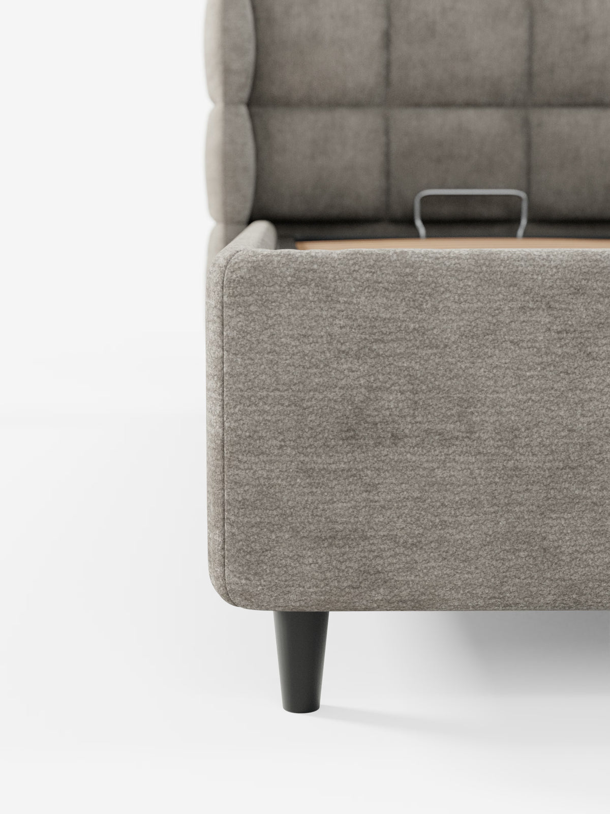 Un primo piano del letto imbottito RAGNAR con contenitore e testiera capitonné avvolgente di Letti.store, rivestito in un elegante tessuto grigio certificato Oeko-Tex® con gambe nere. L’immagine mostra l’angolo inferiore della pediera e della sponda laterale, mettendo in risalto il rivestimento morbido e testurizzato e i caratteristici dettagli capitonné della testiera. Sulla base del letto, su sfondo bianco, è visibile una piccola maniglia in metallo.