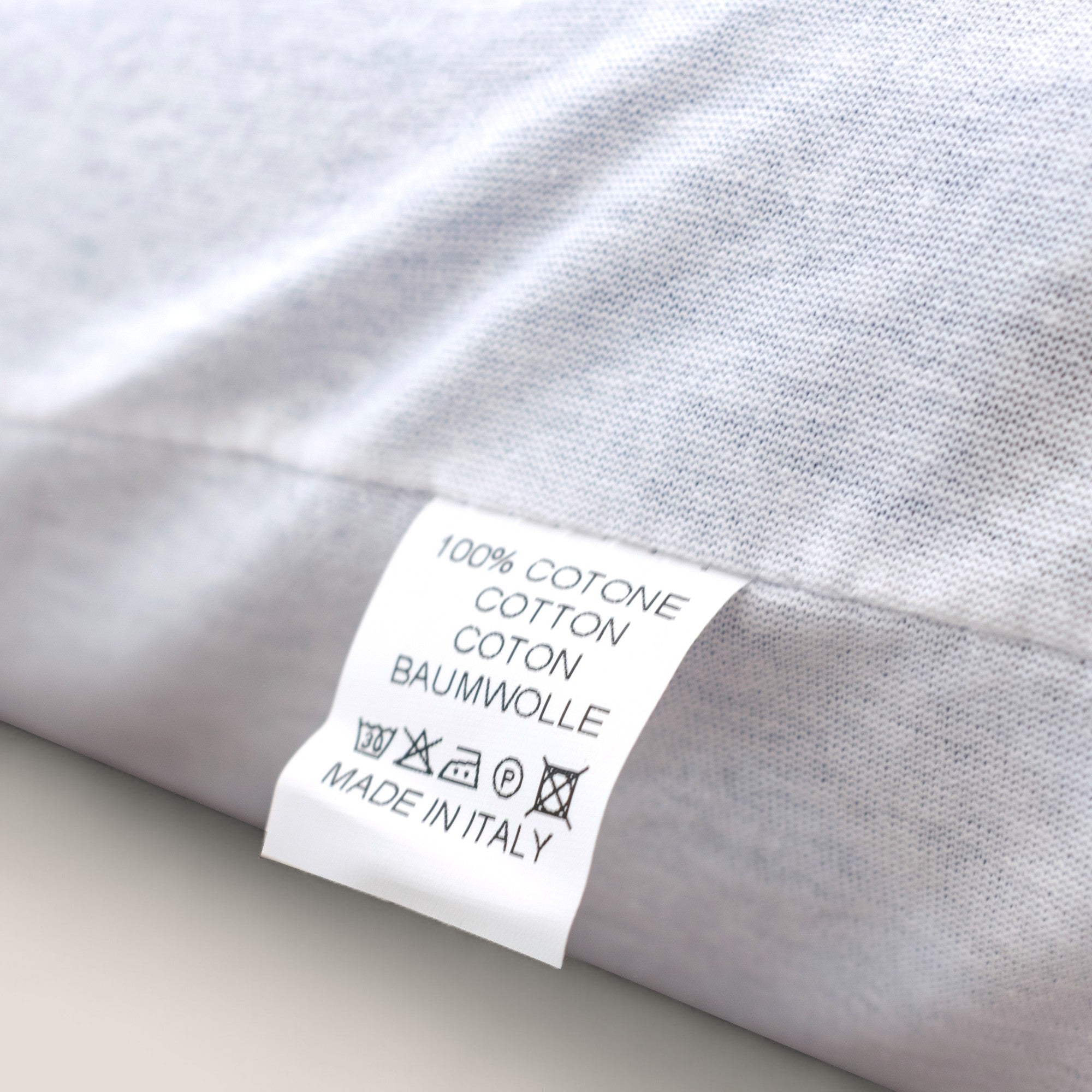 Primo piano di un'etichetta per la cura del tessuto di AERION-BREEZE di Divani.Store, che riporta "100% COTONE" e "MADE IN ITALY". I simboli di lavaggio appaiono in basso, perfetti per il comfort rinfrescante e termoregolatore di questo cuscino in memory gel e per il suo sostegno ergonomico.