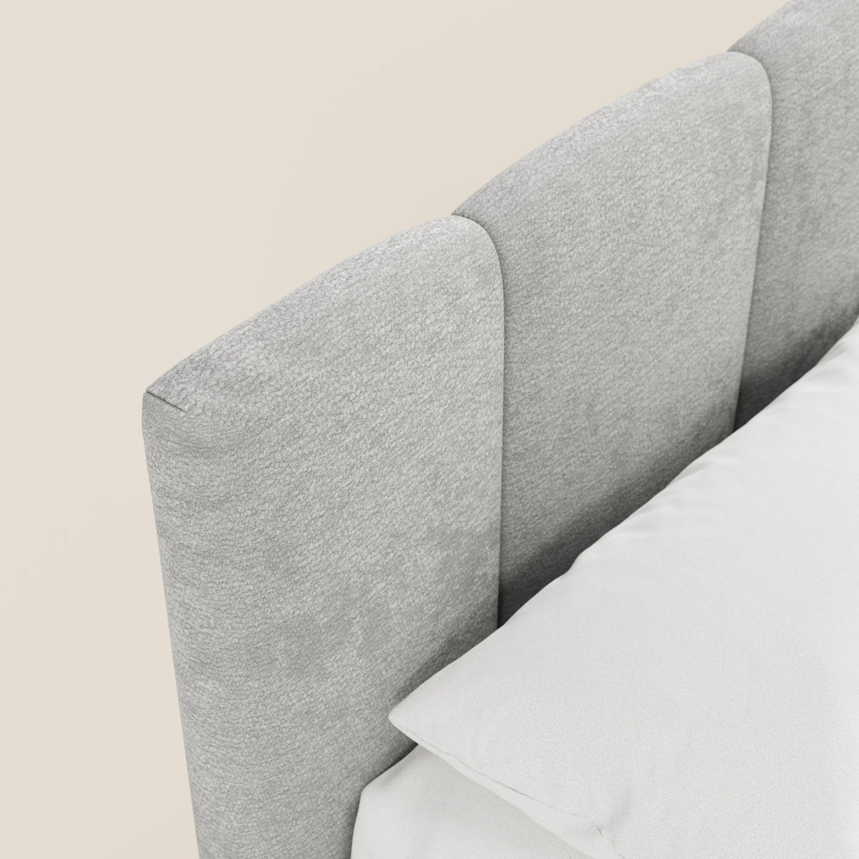 Un primo piano del letto imbottito ARKAM con contenitore di Letti.store mette in risalto la sua testiera in tessuto grigio chiaro, in stile shabby chic con pannelli verticali e certificata Oeko-Tex®, con un cuscino e un lenzuolo bianchi su uno sfondo bianco sporco.