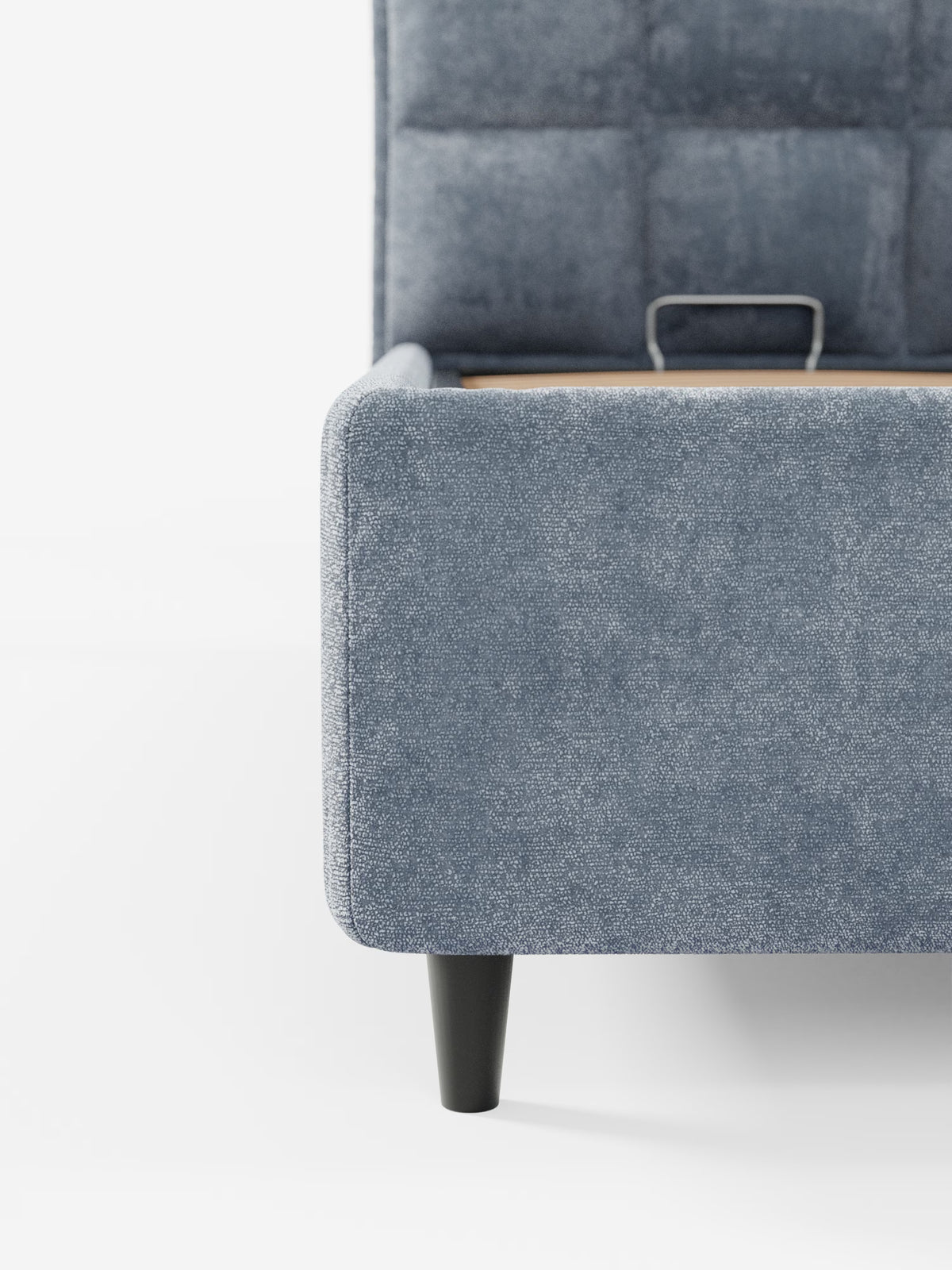 Primo piano del letto imbottito con contenitore AURO di Letti.store, che mette in risalto un angolo rivestito in morbido tessuto chenille blu-grigio certificato Oeko-Tex®. Il letto presenta un’elegante testiera alta capitonné e un pannello laterale dalle linee morbide e curve. Una singola gamba affusolata nera sostiene la base, dove una piccola maniglia in metallo è fissata al telaio in legno. Lo sfondo è bianco a tinta unita.