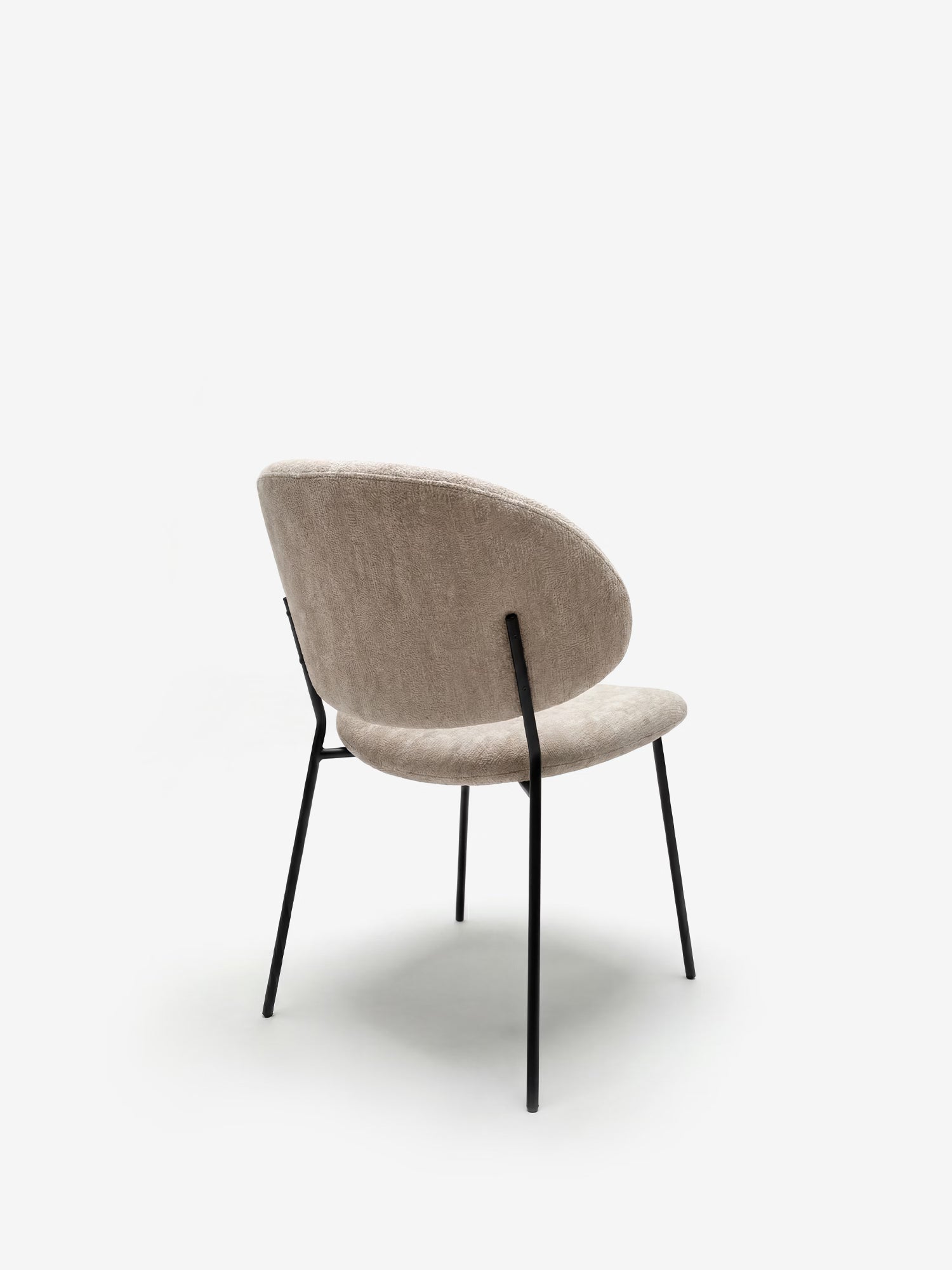 La sedia Bruma di Divani.Store presenta un design minimalista con schienale avvolgente in tessuto goffrato beige chiaro e quattro gambe sottili in metallo nero, leggermente inclinate verso l’esterno; è stata fotografata da dietro su uno sfondo bianco.