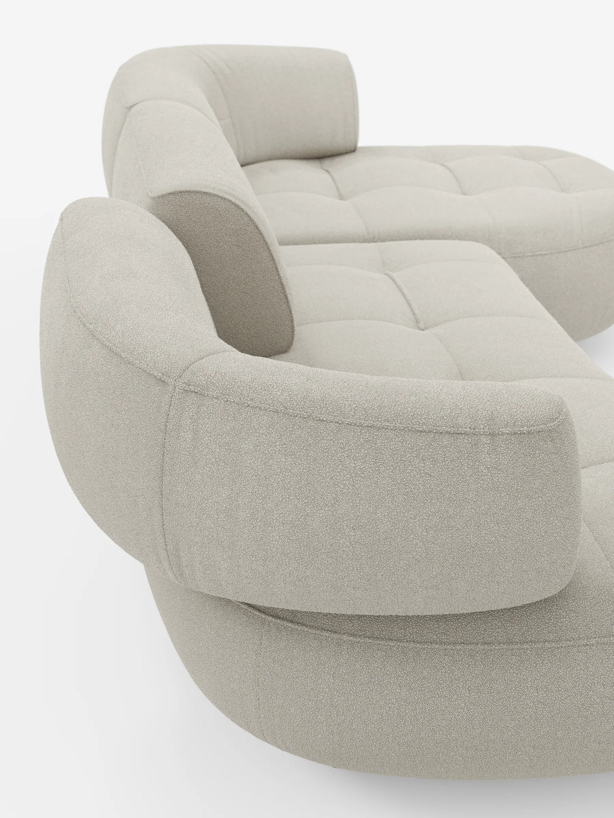 Il divano angolare Croissant, di colore beige chiaro, dal design a fagiolo e con schienali scorrevoli in morbido tessuto effetto cotone smacchiabile T33, presenta una seduta trapuntata e schienali arrotondati su uno sfondo bianco a tinta unita.