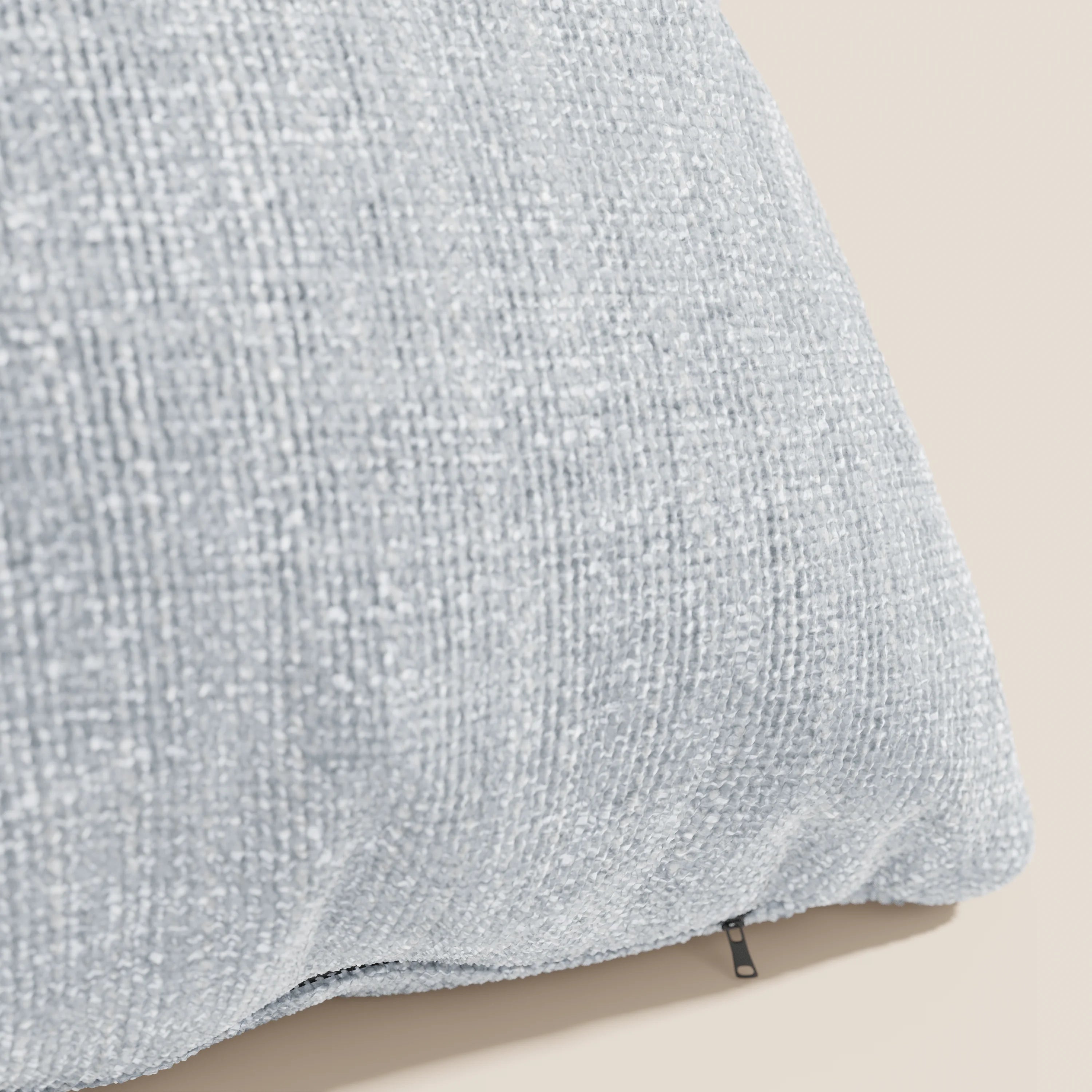 Primo piano del cuscino Lyon 45x45 cm in T06 ghiaccio di Divani.Store, caratterizzato da un tessuto morbido intrecciato impermeabile e da una texture sottile, su uno sfondo beige con una piccola cerniera nera sul bordo inferiore.
