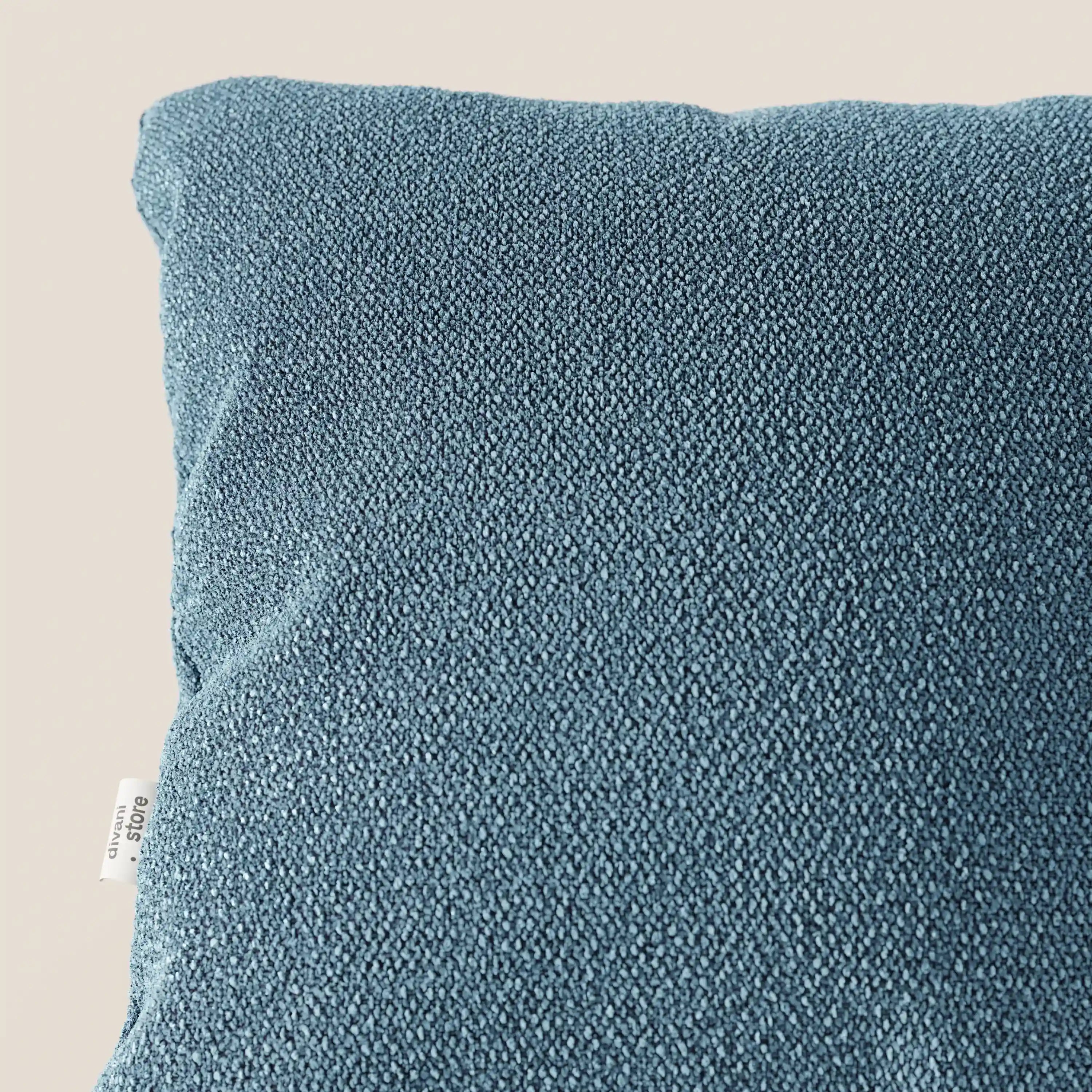 Primo piano del cuscino Lambada di Divani.Store in blu, realizzato in un tessuto fresco, antimacchia e impermeabile simile al cotone, caratterizzato da una piccola etichetta laterale bianca con testo nero e da una trama visibile su uno sfondo neutro.