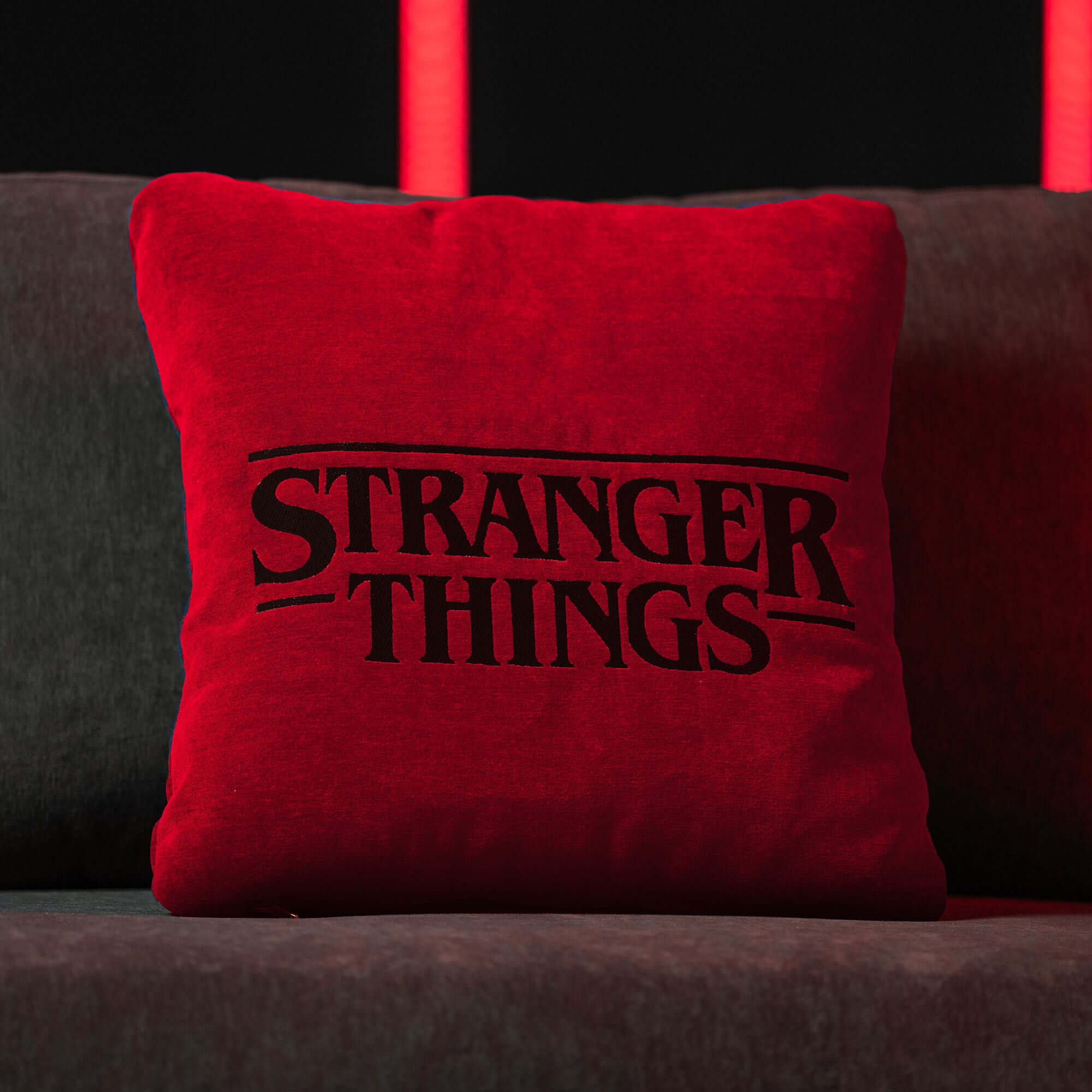Un centrotavola di grande effetto, il Cuscino Stranger Things Limited Edition con Ricamo 45x45 rosso di Divani.Store è caratterizzato da un audace logo nero e da un tessuto rosso, che risalta su un divano grigio scuro contro le luci al neon, con un materiale facile da pulire.