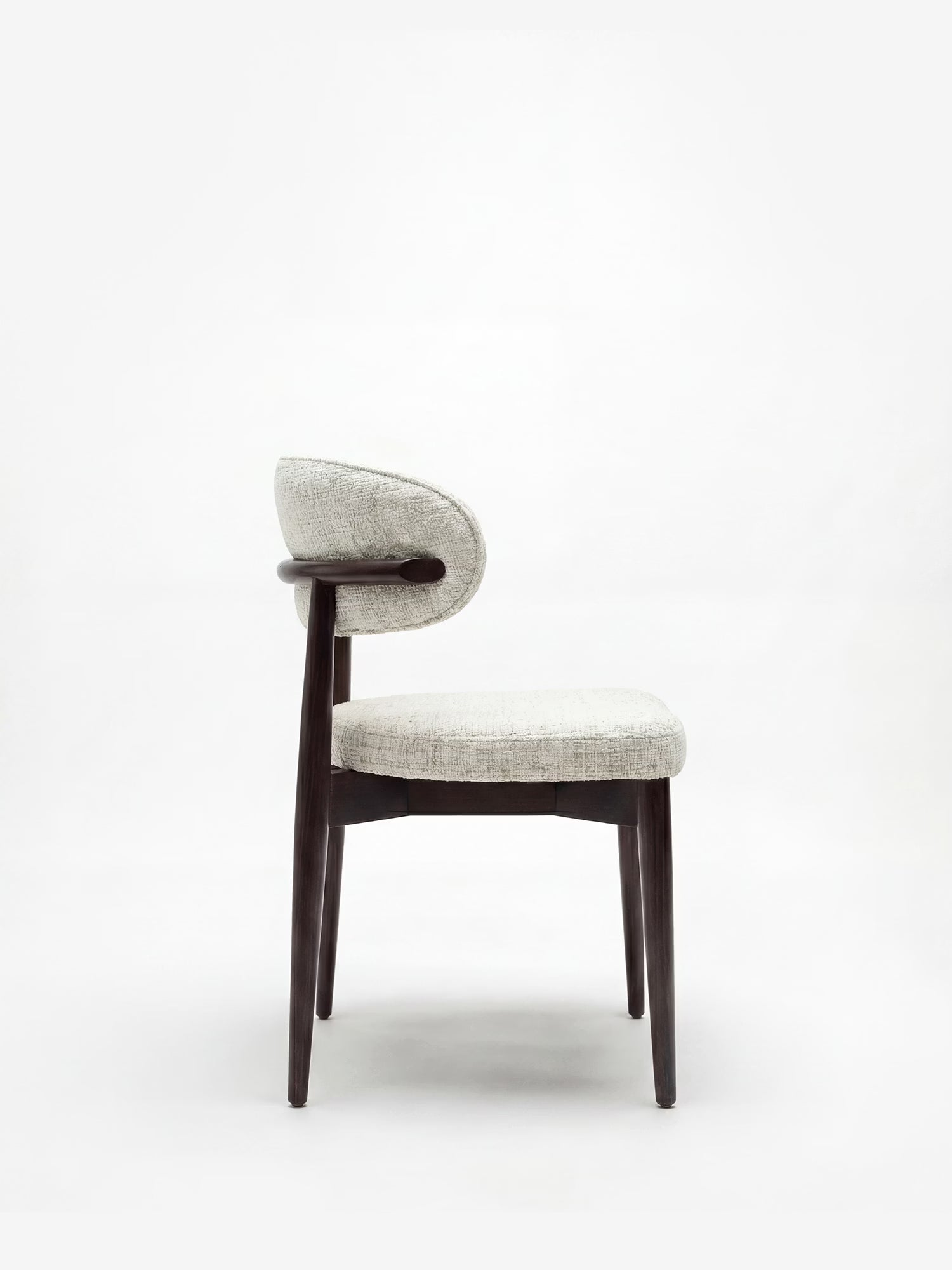 La sedia Dafne di Divani.Store presenta una struttura e gambe in legno massello wengé, uno schienale rotondo imbottito e una seduta in tessuto ciniglia certificato OEKO-TEX® dalla trama leggera, qui raffigurata di profilo su uno sfondo bianco a tinta unita.