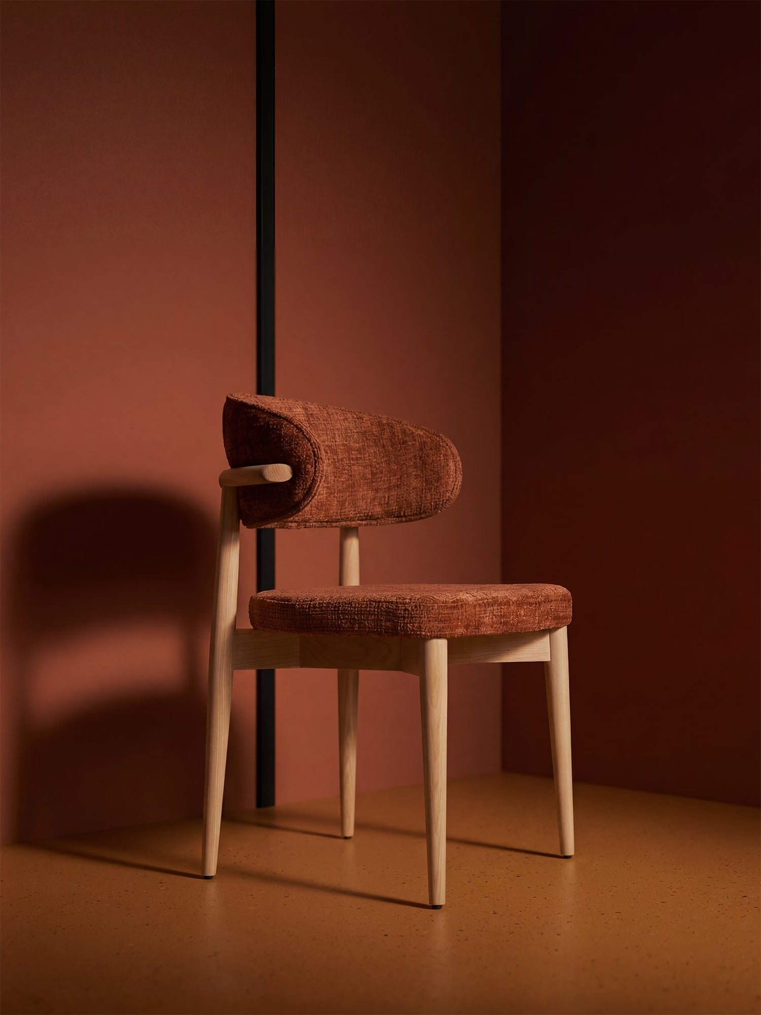 La sedia imbottita Dafne di Divani.Store, con gambe in rovere massello naturale e tessuto in ciniglia certificato OEKO-TEX® color ruggine, presenta uno schienale ricurvo e proietta un’ombra decisa sulle calde pareti marroni e sul pavimento color senape di una stanza minimalista.